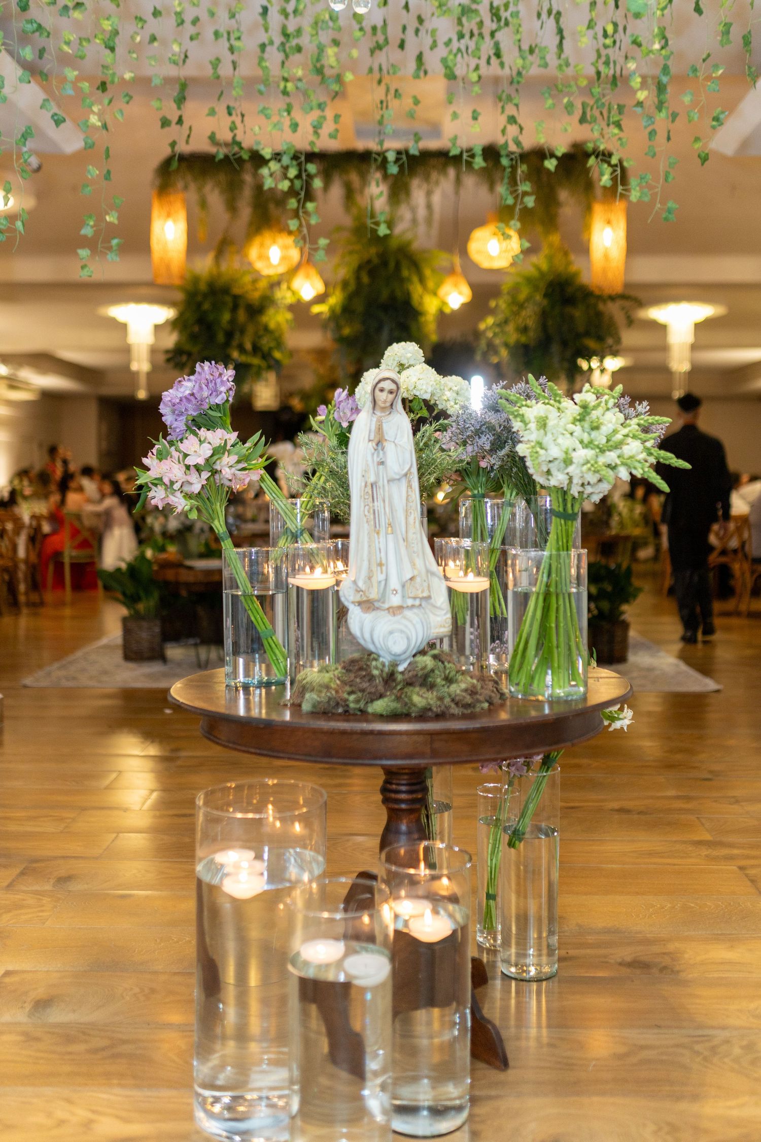 Elegant wedding reception centerpiece featuring a Virgin Mary statue, candles, and fresh flowers on a wooden table.