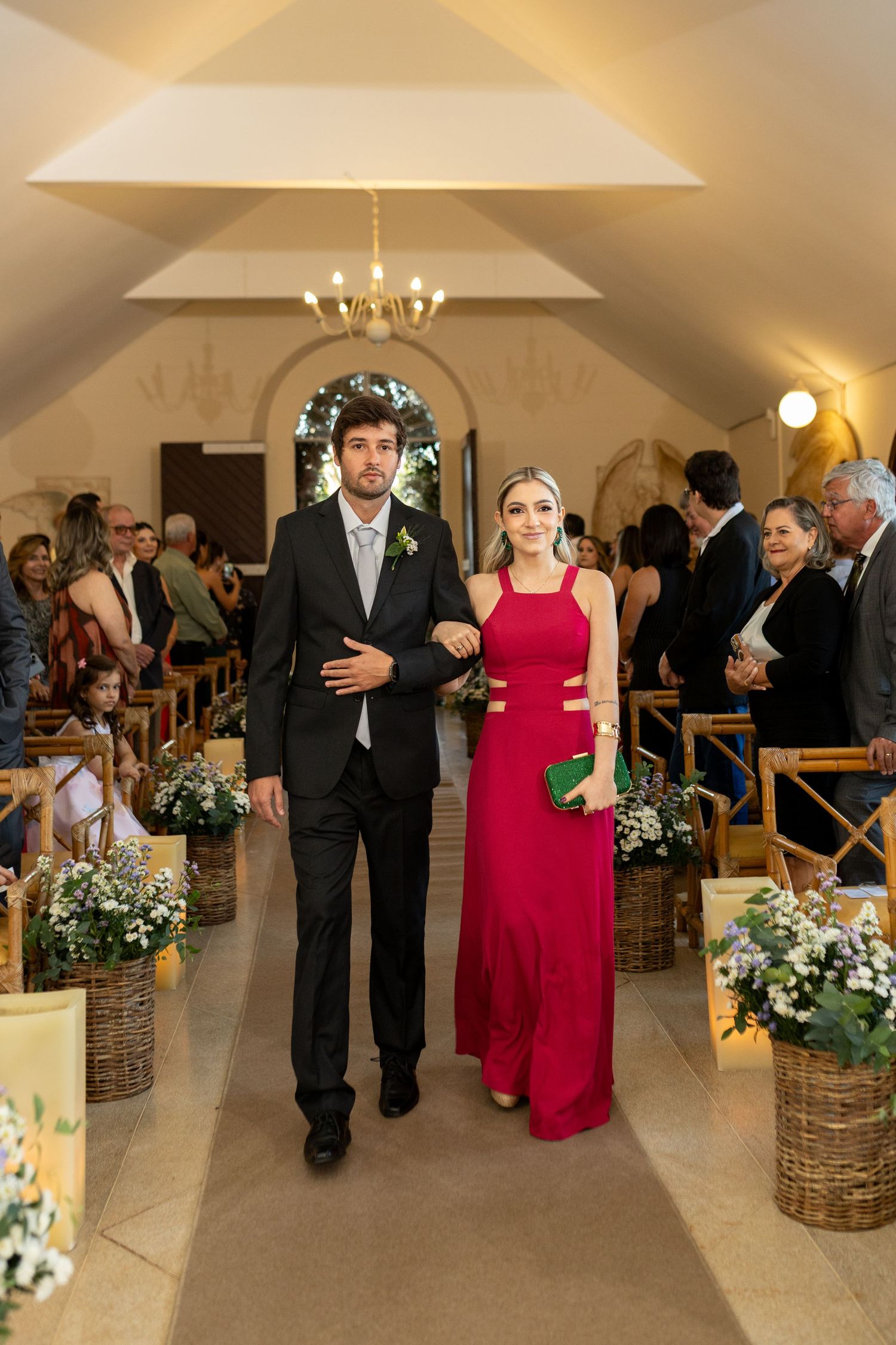 Couple walking down wedding aisle, man in black suit, woman in red gown, floral decorations, chapel setting.