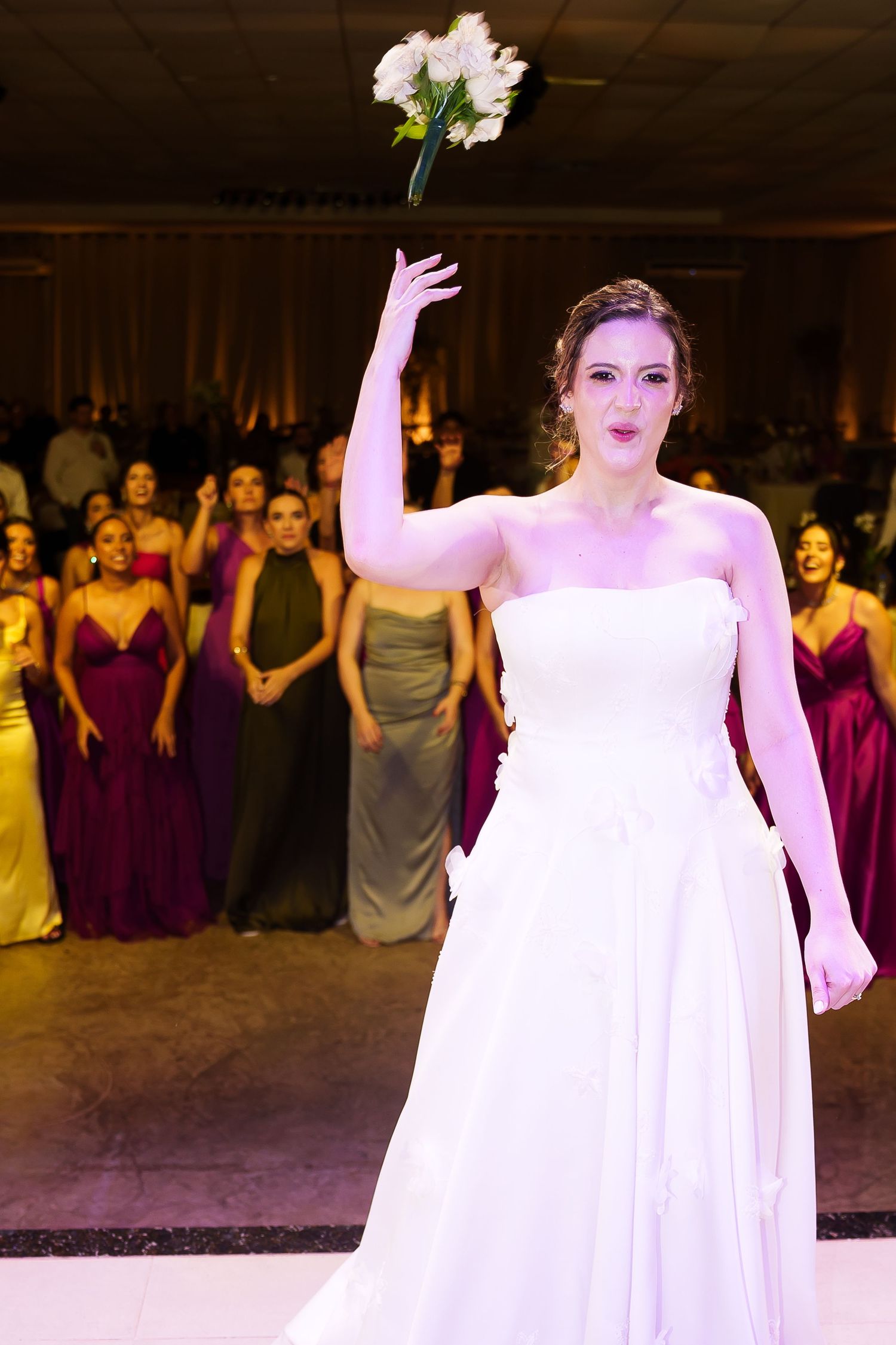Bride in white strapless gown tossing bouquet at wedding reception with bridesmaids in colorful dresses watching.