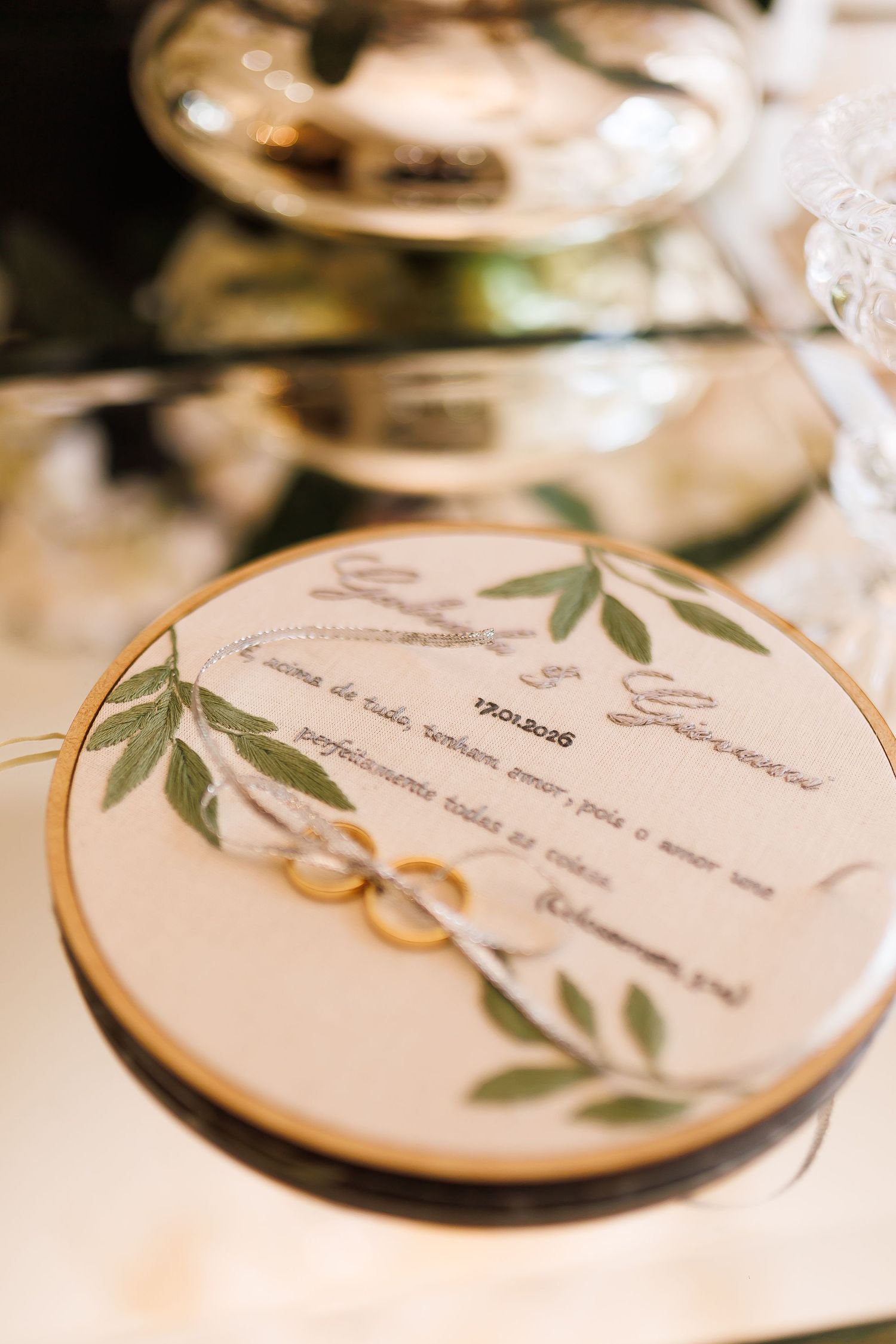 Wedding rings resting on embroidered hoop with green leaves and couple names, elegant bridal detail photography.