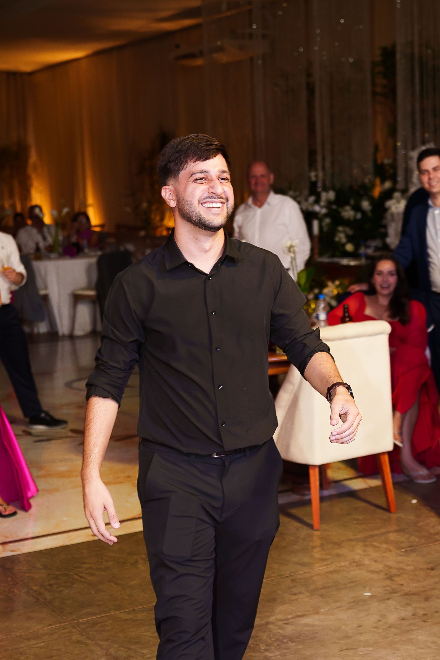 A smiling young man in all-black attire walks confidently on a dance floor at an elegant evening event.