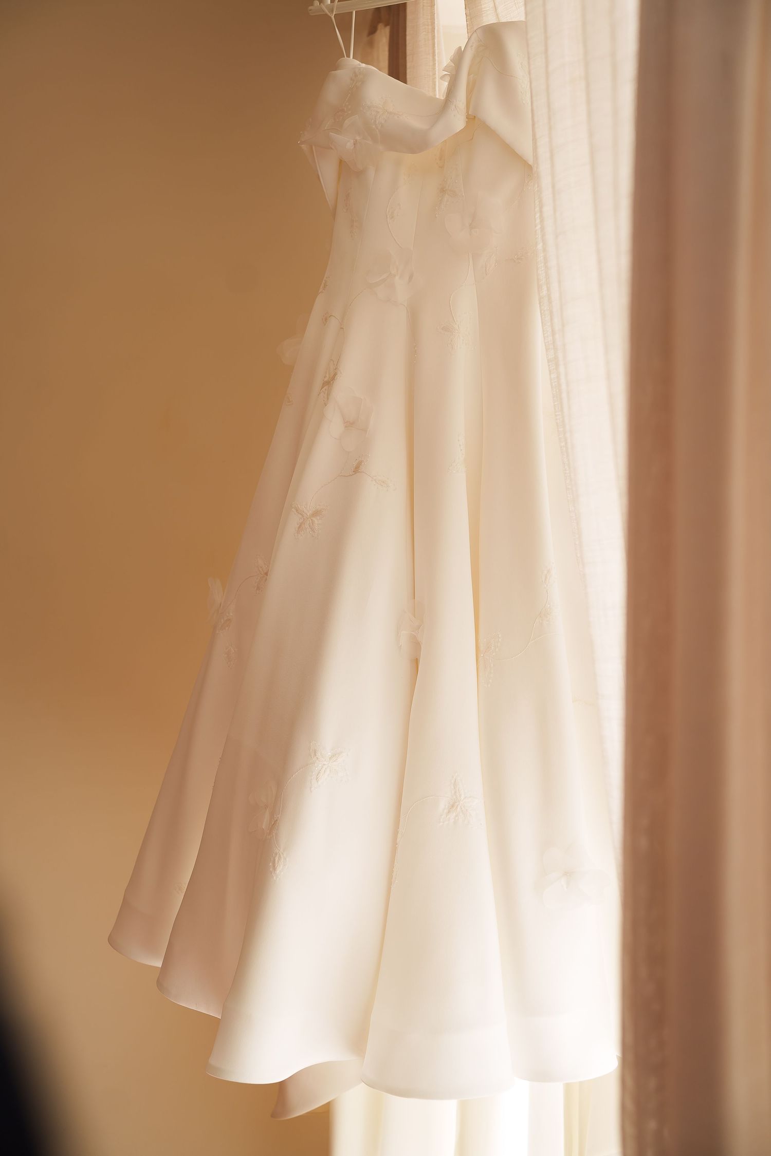 A delicate white wedding dress with floral embroidery hangs elegantly near a sunlit window.