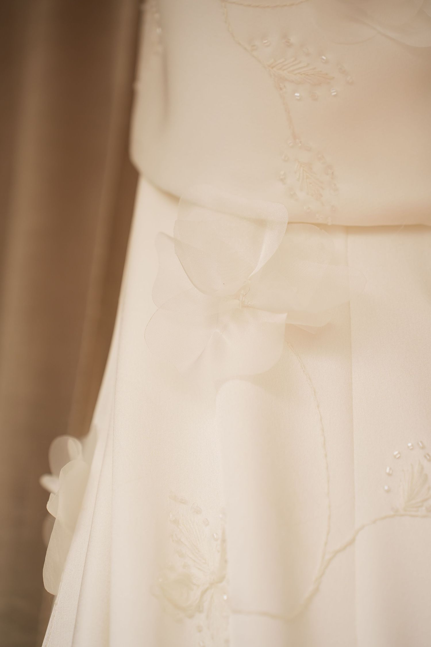 Close-up of an ivory wedding dress with delicate floral embroidery, pearl beading, and a satin ribbon bow at the waist.