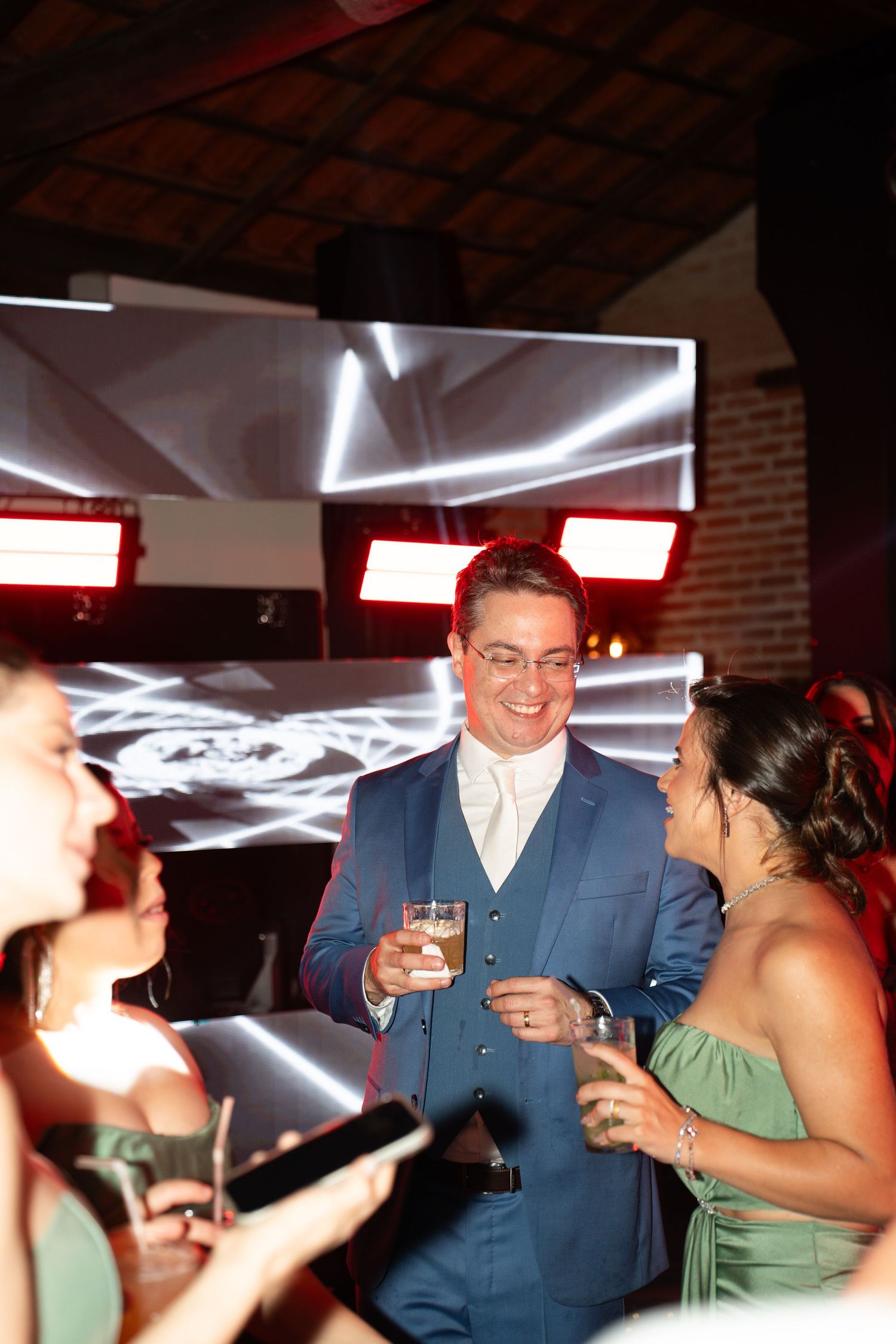 Man in blue suit smiling and holding a drink at an elegant evening celebration with guests in formal attire.