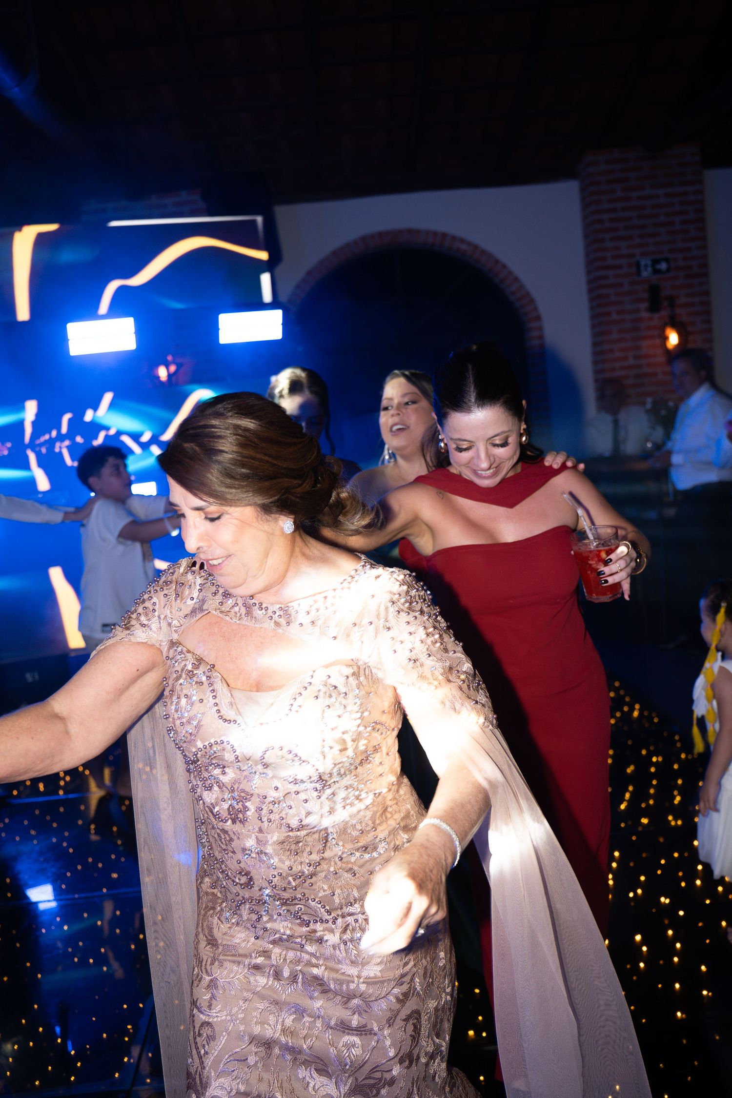 Women in elegant gowns dancing joyfully at a formal evening event with blue lighting.