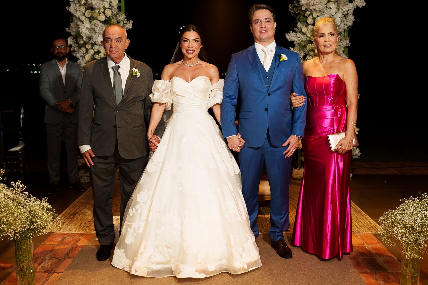 Bride in white gown poses with groom in blue suit and family members in formal attire at elegant wedding ceremony.