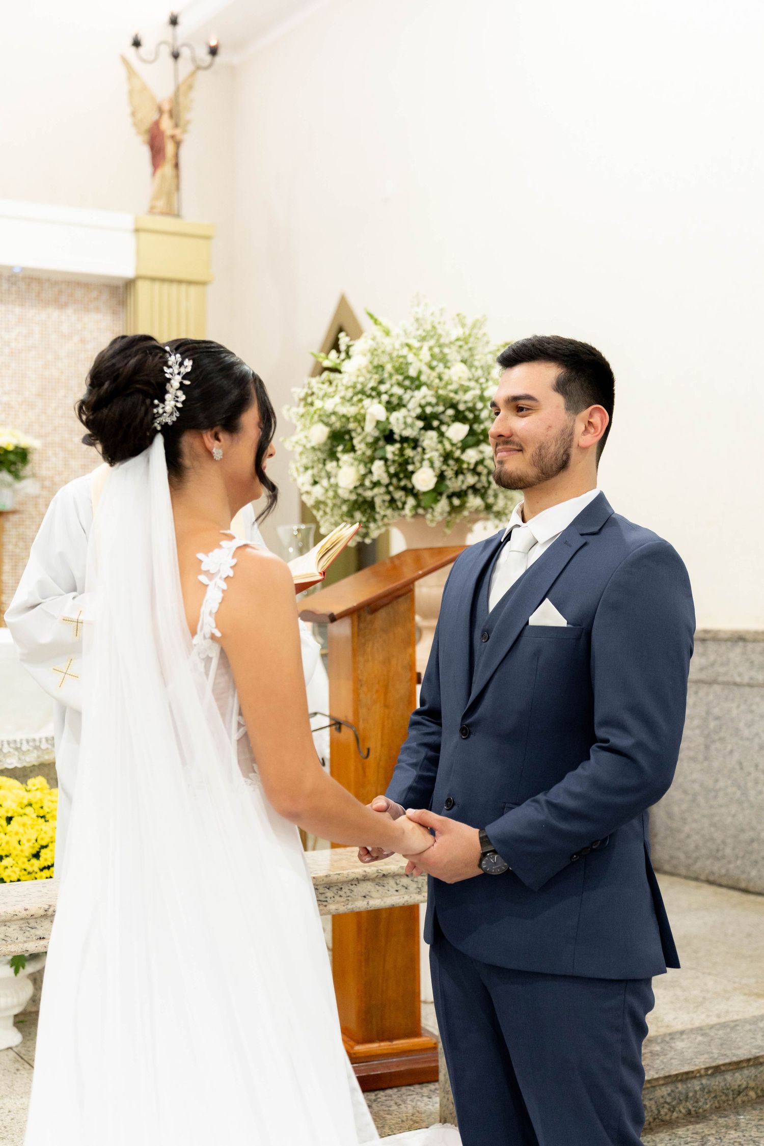 Bride and groom holding hands at church altar, bride in white gown with veil, groom in navy suit.