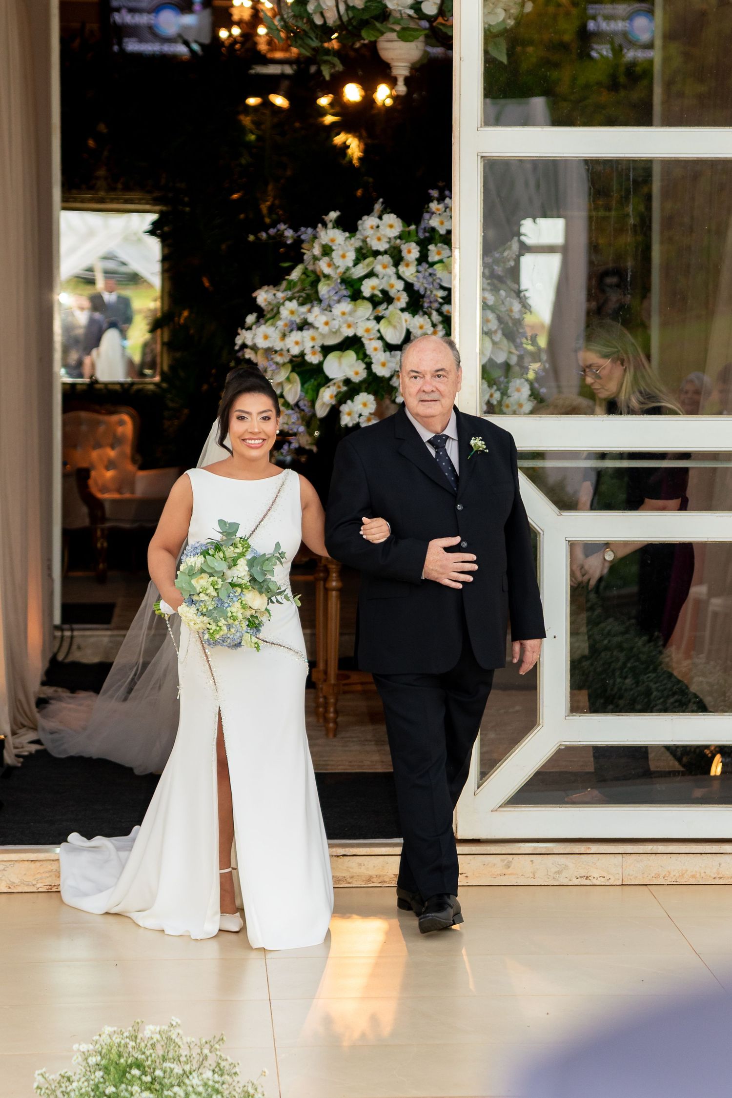 Bride in white gown walks down aisle with older man in black suit, white floral arrangements in background.