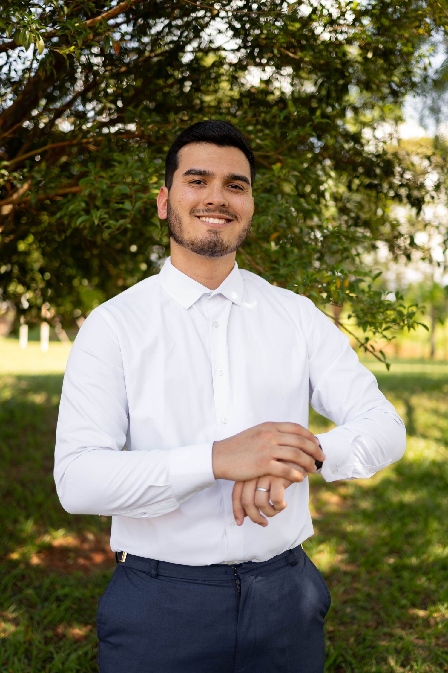 Smiling young man in white dress shirt adjusting cufflinks outdoors with green trees in background.