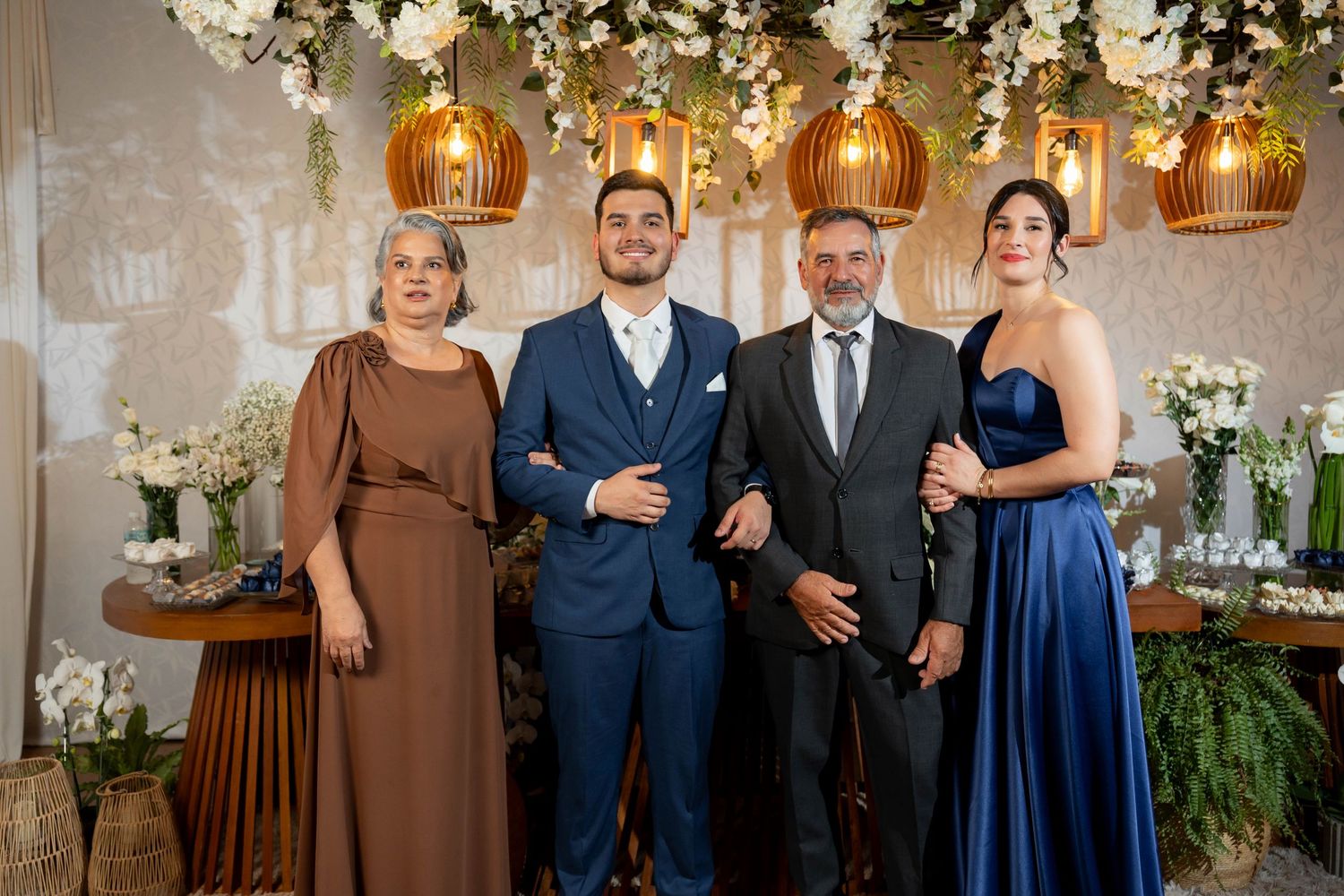 Four elegantly dressed people posing together at a wedding reception with floral decor and golden pendant lights.