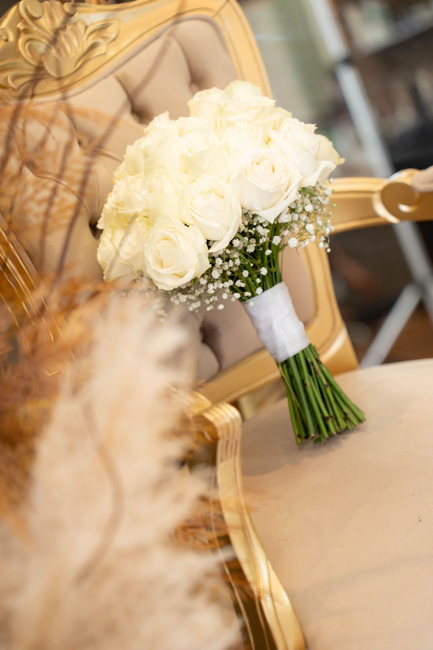 White rose bridal bouquet with baby's breath tied with white ribbon resting on a gold ornate chair.
