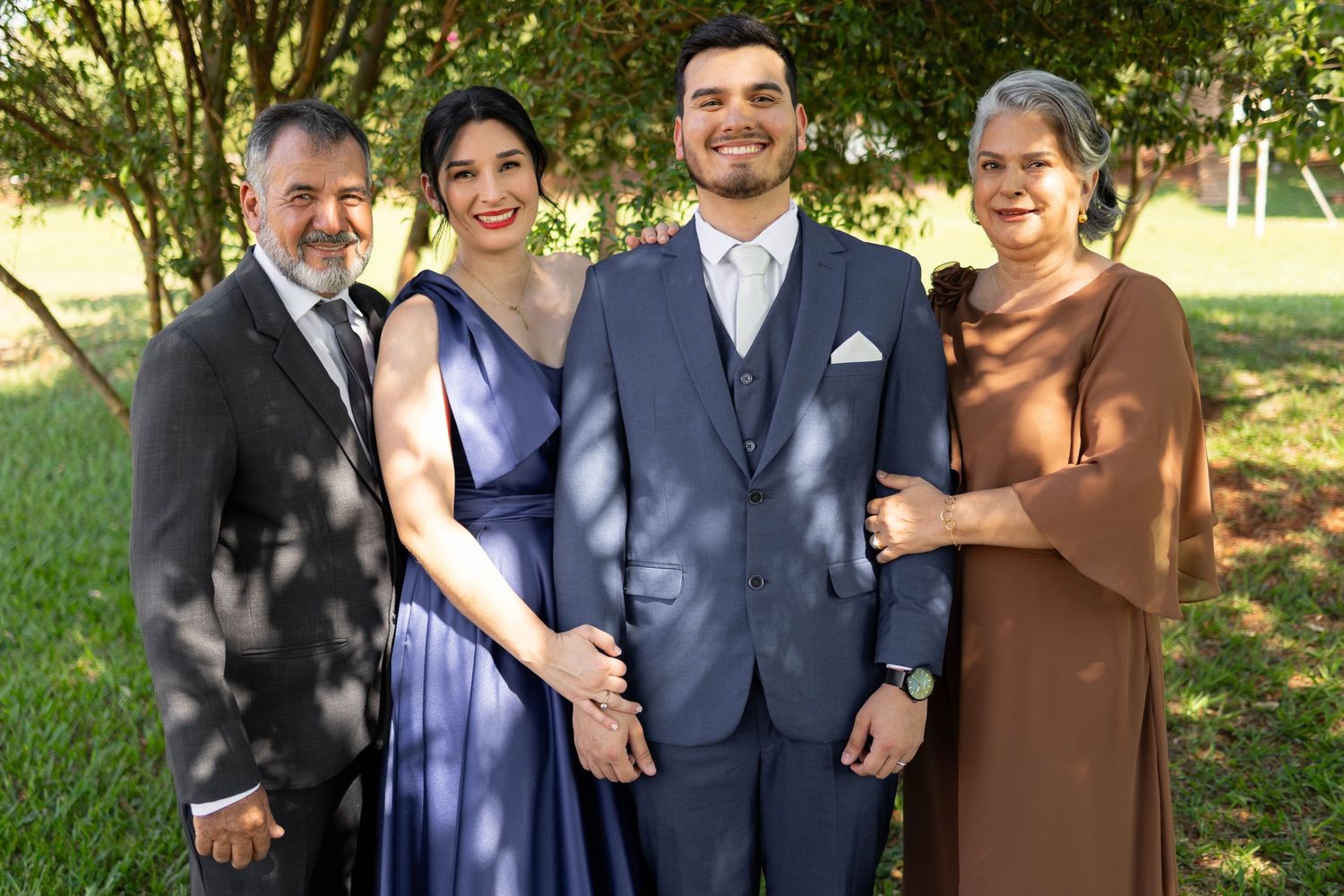 Four elegantly dressed people posing together outdoors at a formal event, smiling in a lush green garden setting.
