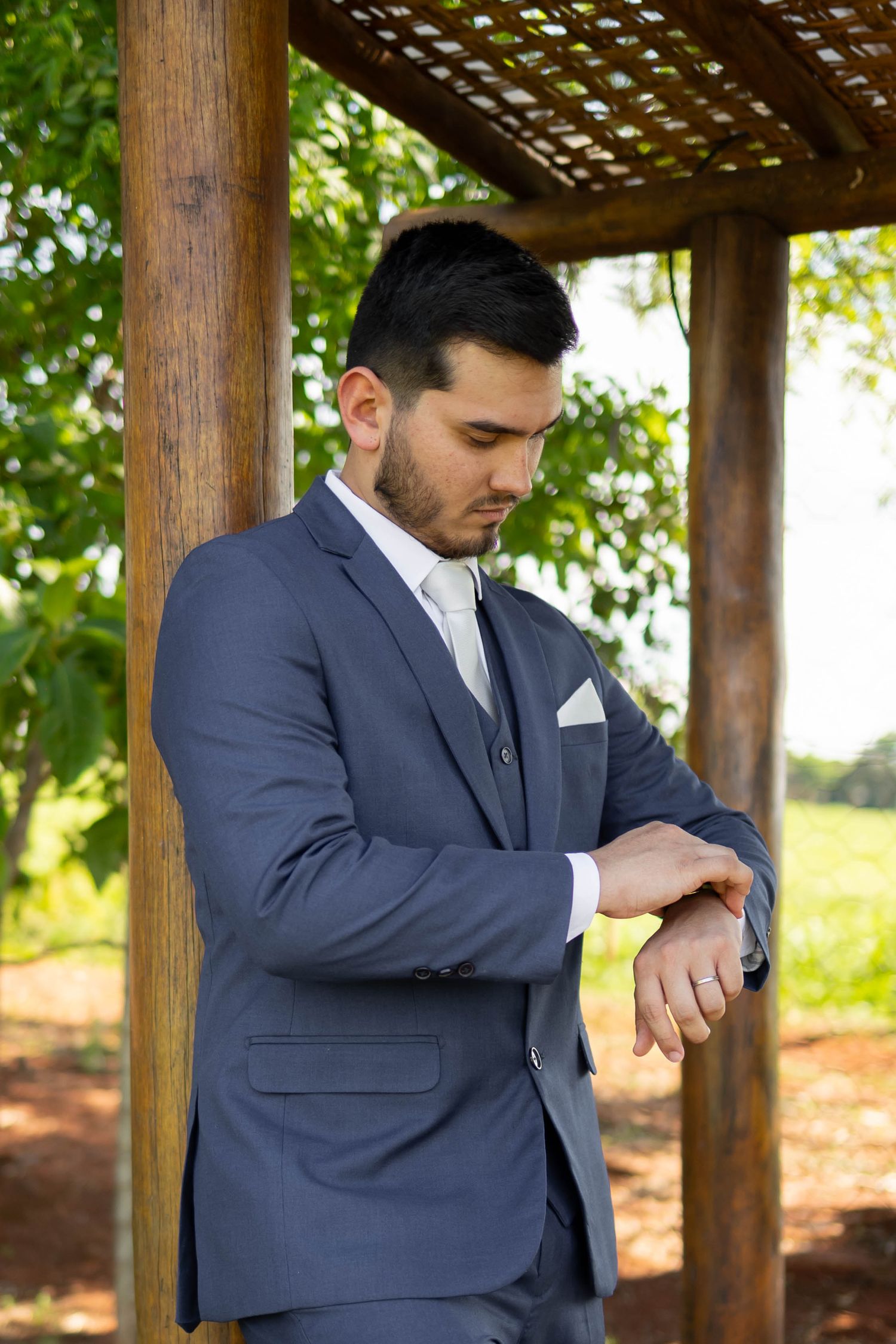 A well-dressed young man in a blue suit checks his watch under a wooden pergola surrounded by lush green trees.
