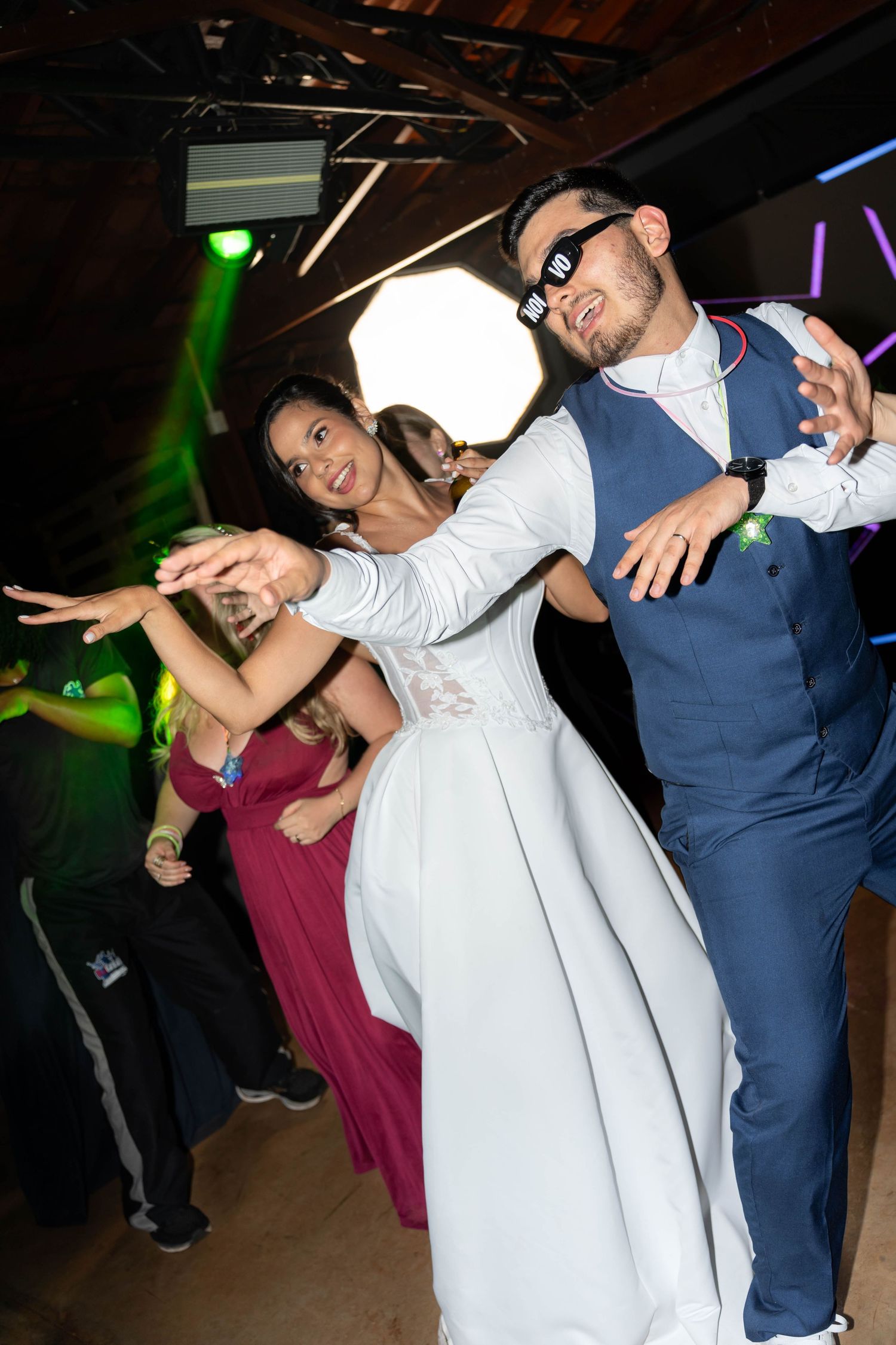 Bride and groom dancing energetically at wedding reception, groom wearing sunglasses and blue suit, bride in white gown.