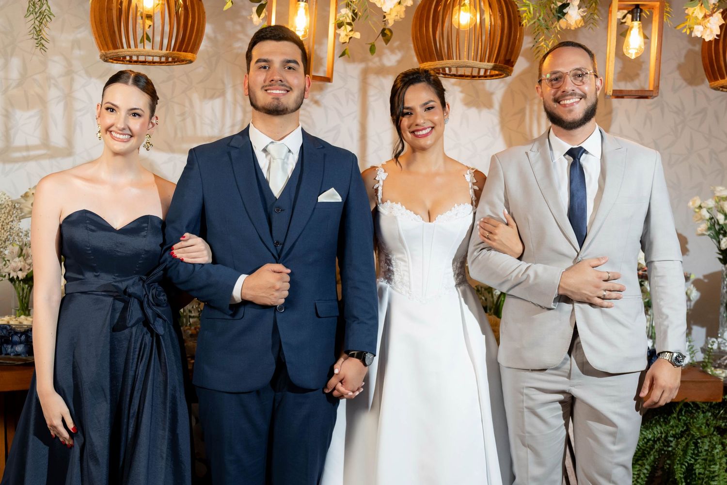 Wedding party of four posing indoors, bride in white gown, groom in navy suit, guests in formal attire, floral decor background.