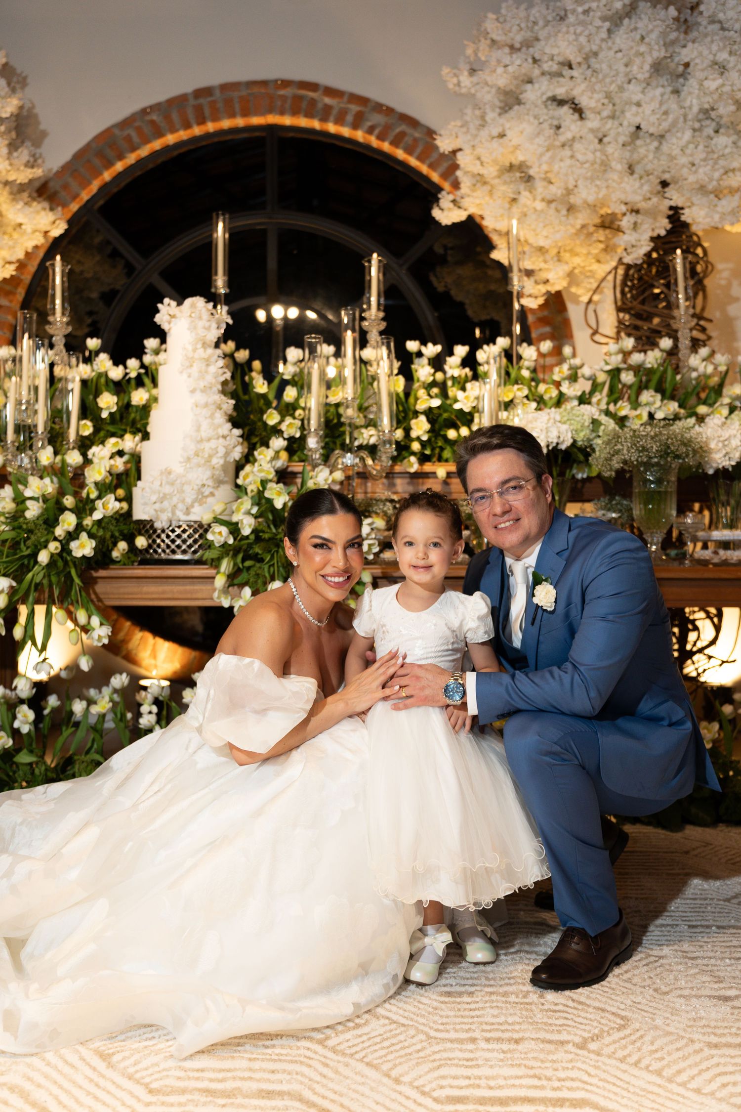 Bride and groom in blue suit pose with young flower girl before floral wedding backdrop with white blooms and candles.
