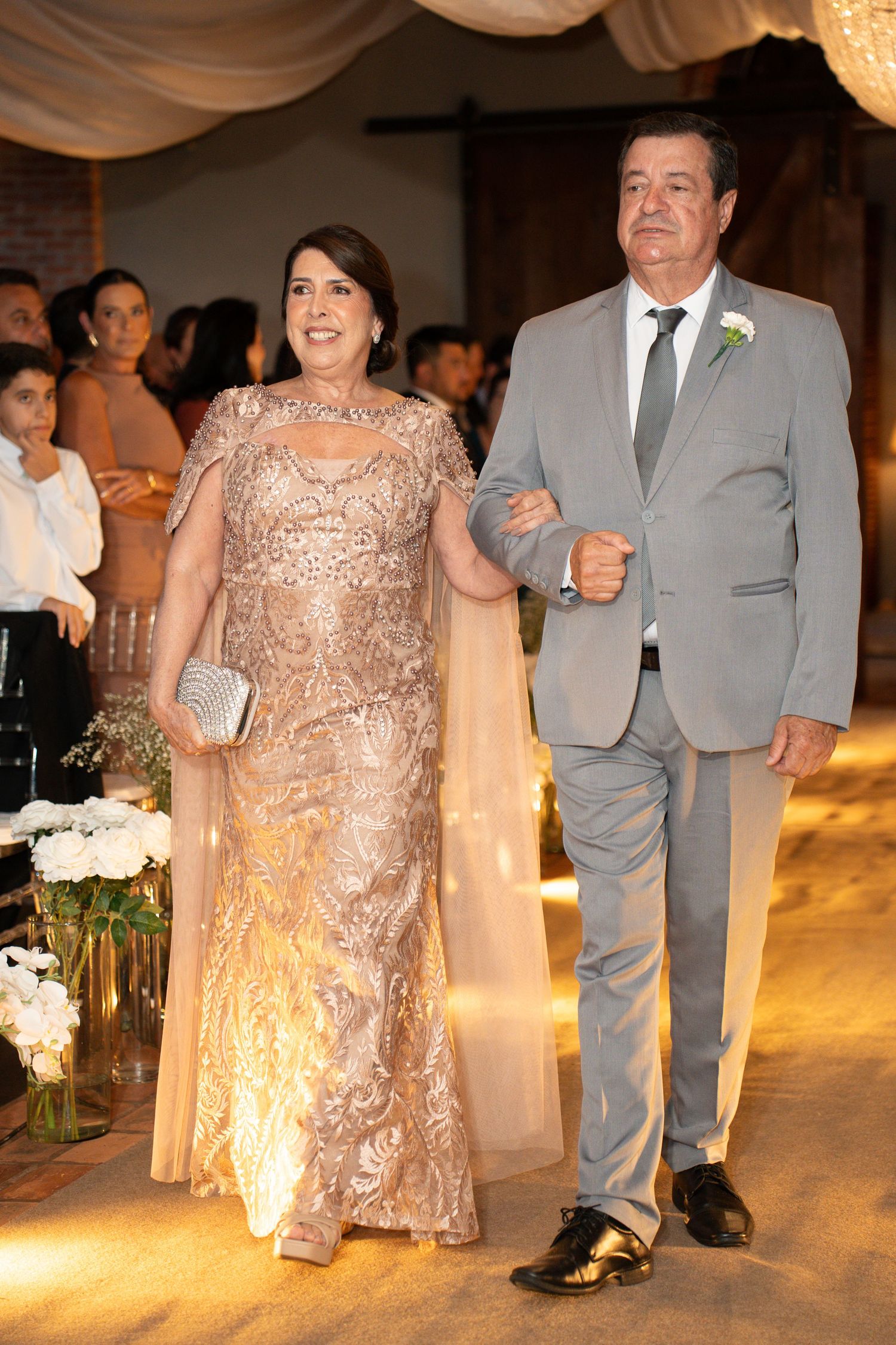 Elegant couple walking down wedding aisle, woman in gold lace gown, man in gray suit with boutonniere.