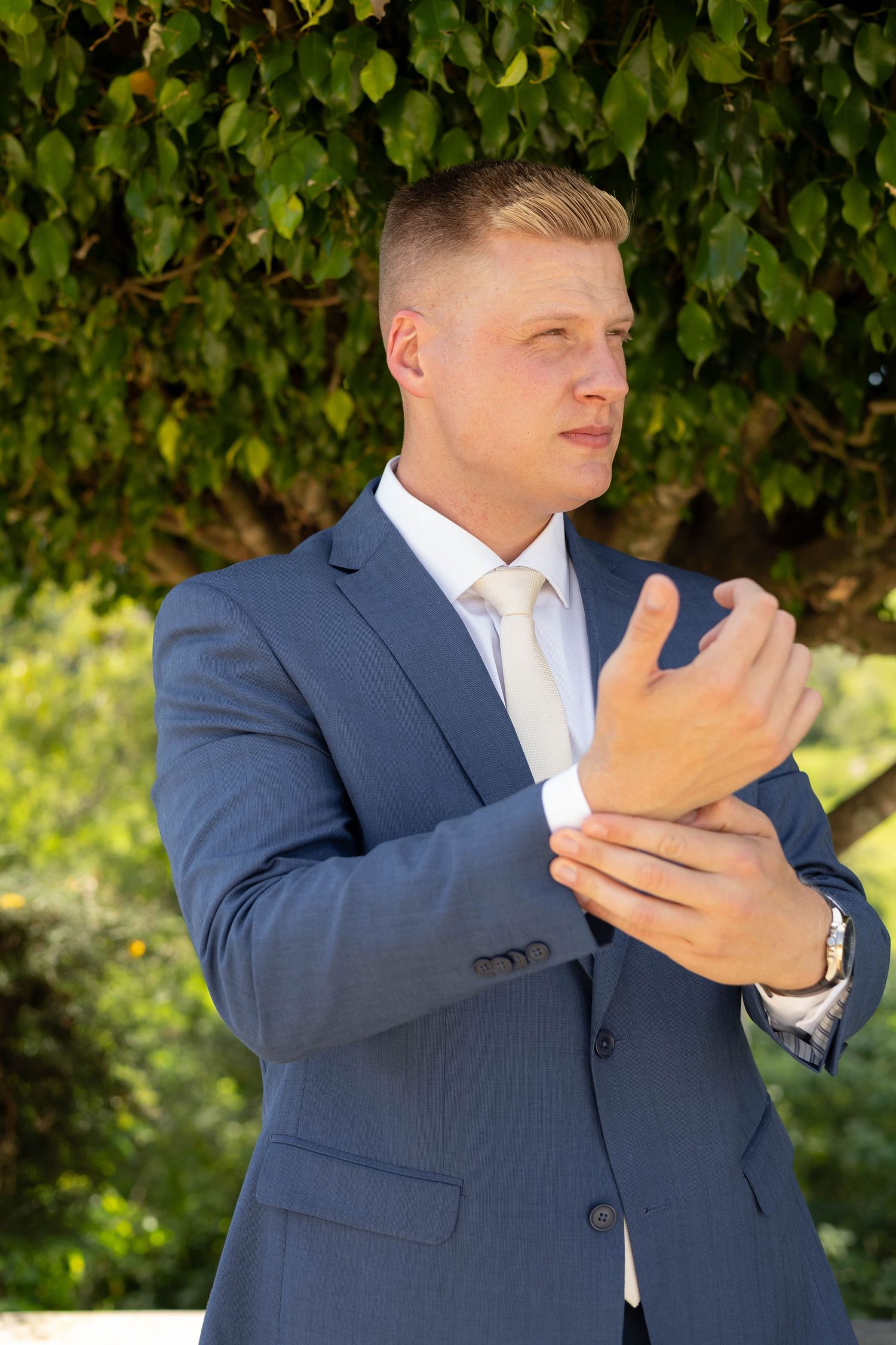 Young man in blue suit adjusting cufflinks outdoors with green trees in background.