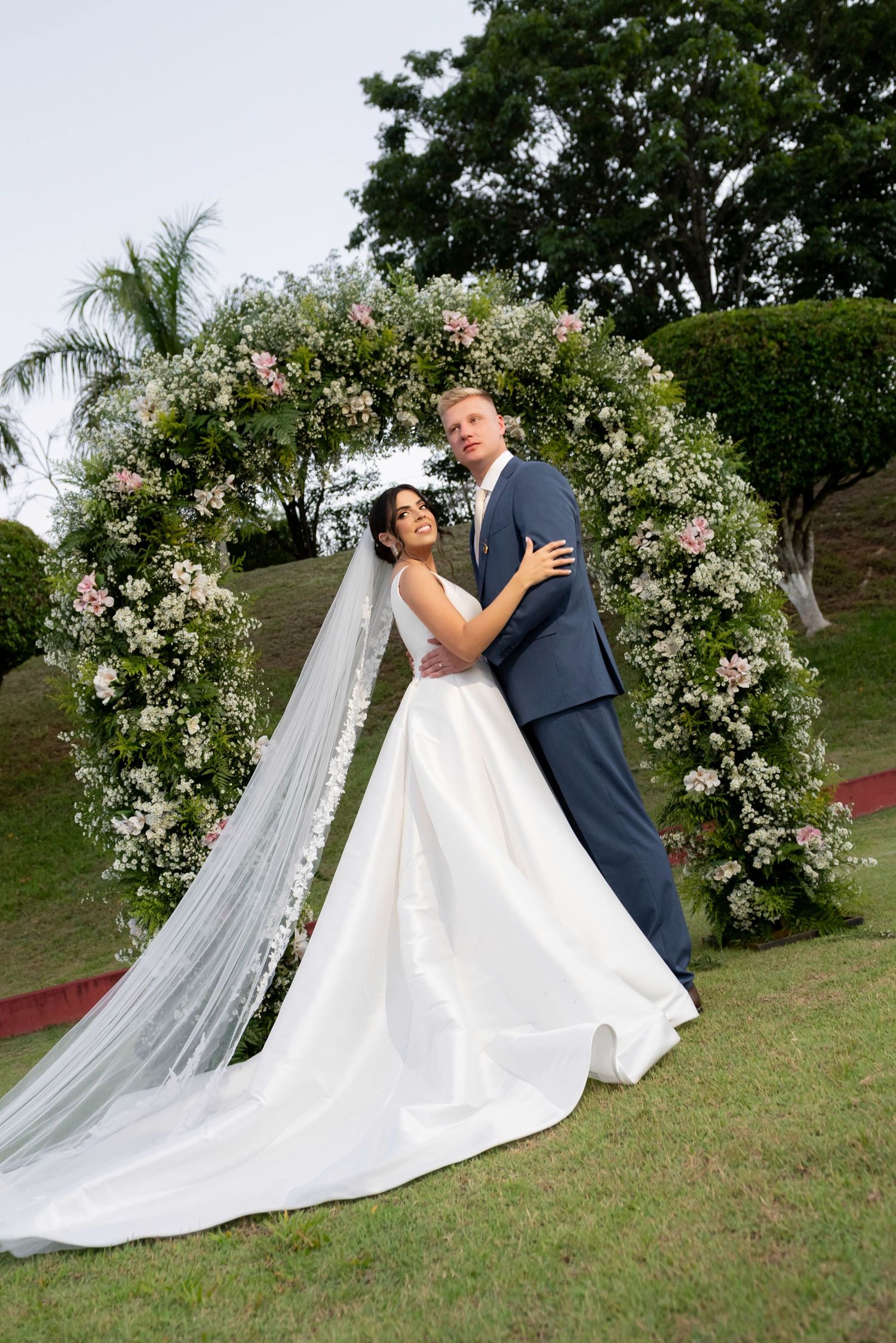 Bride and groom embracing under a floral arch outdoors, bride in white gown with veil, groom in gray suit.
