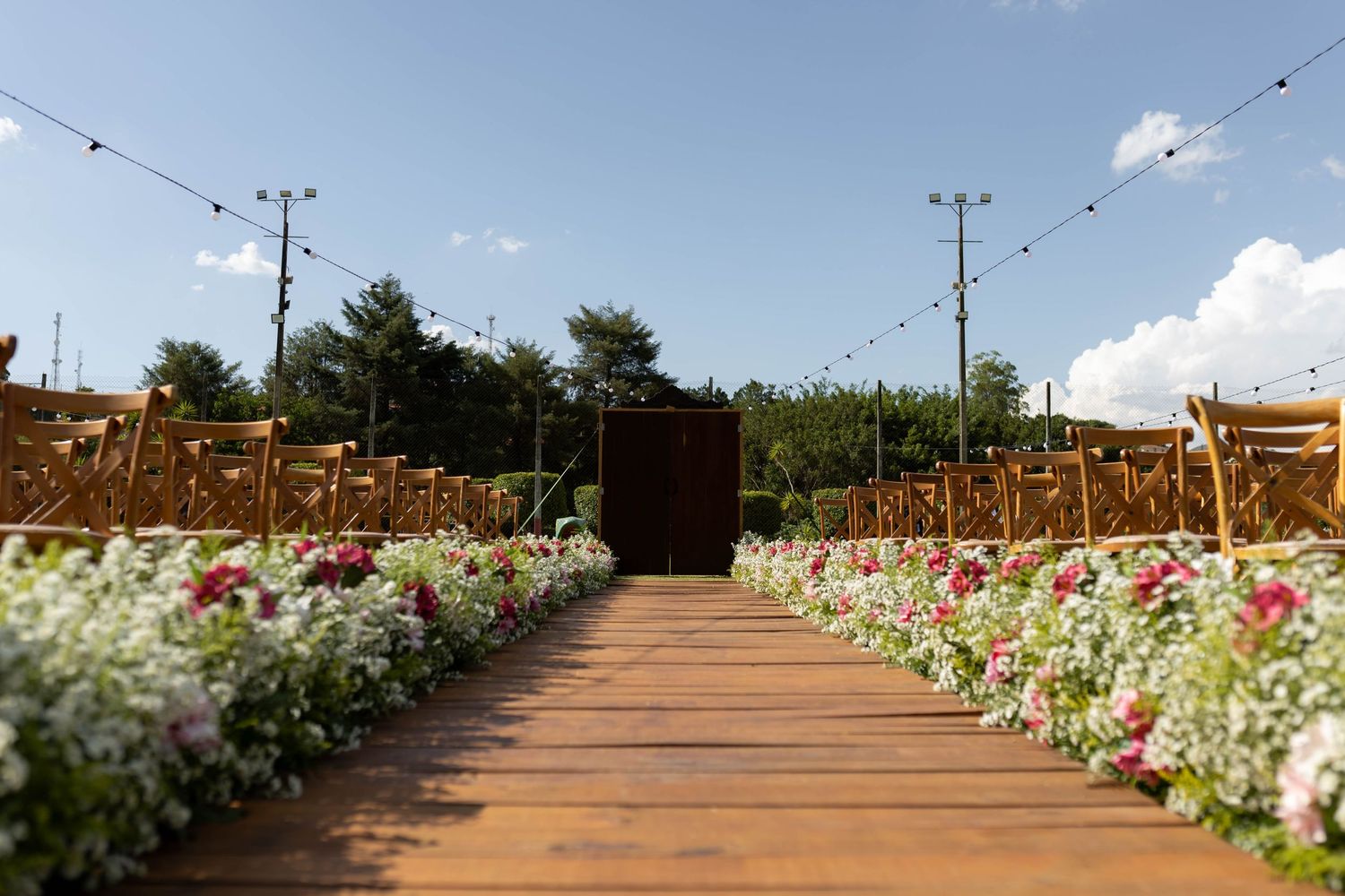 Wooden aisle lined with colorful flowers and golden chairs leads to a rustic door under a clear blue sky with string lights.