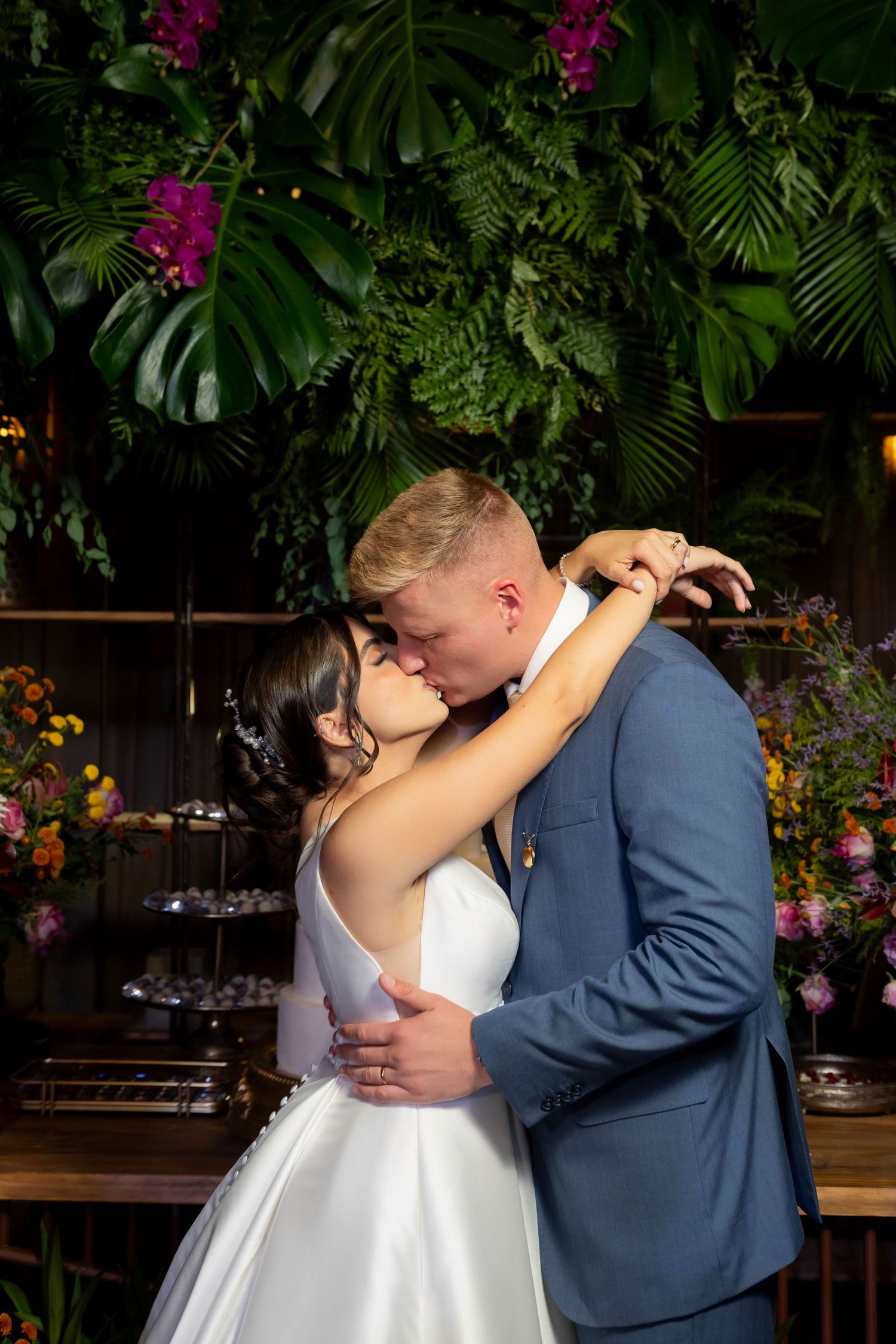 Bride and groom share a romantic kiss at their wedding reception surrounded by lush tropical greenery and colorful flowers.