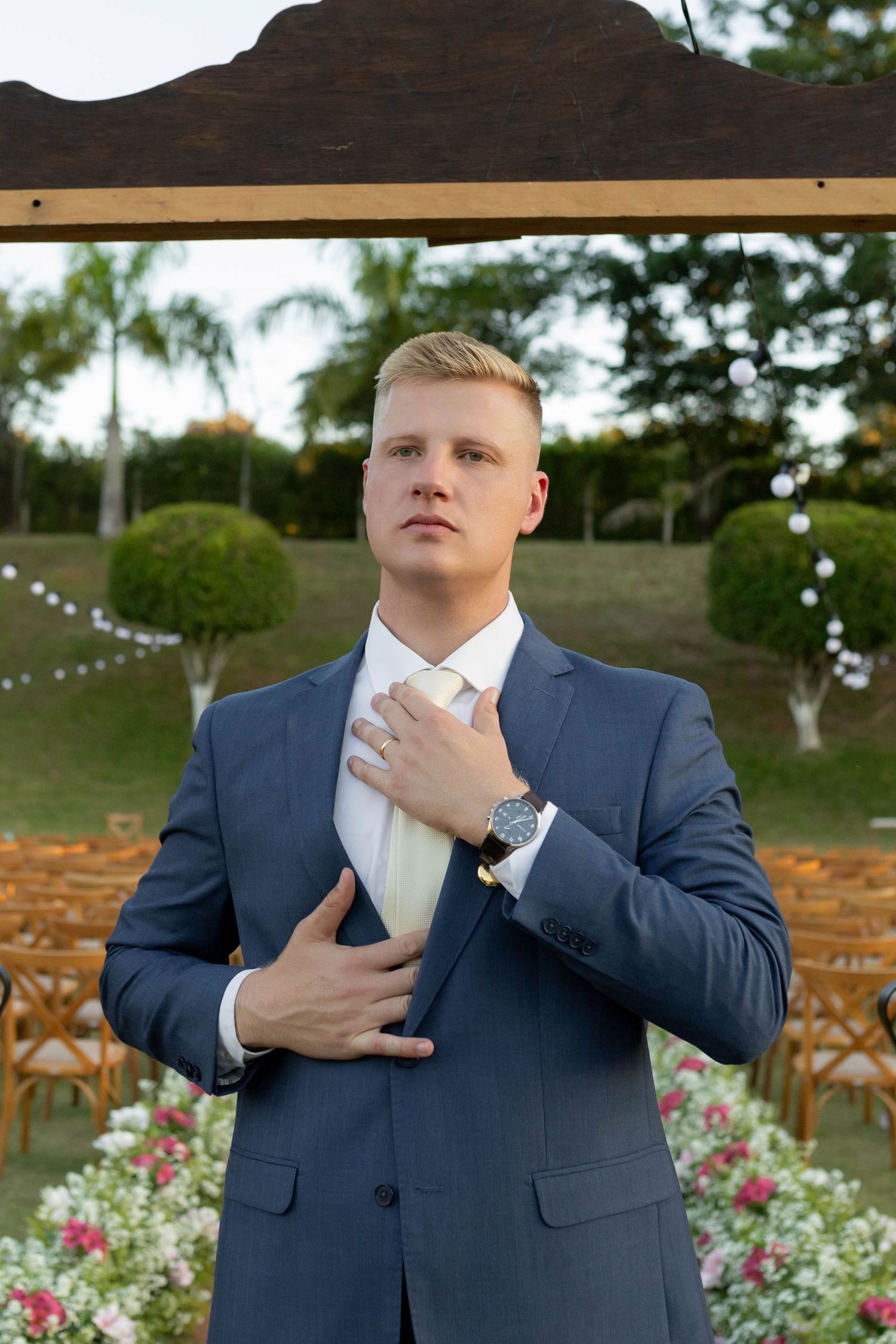 Groom in navy suit adjusting his tie at an outdoor wedding ceremony with floral arrangements.