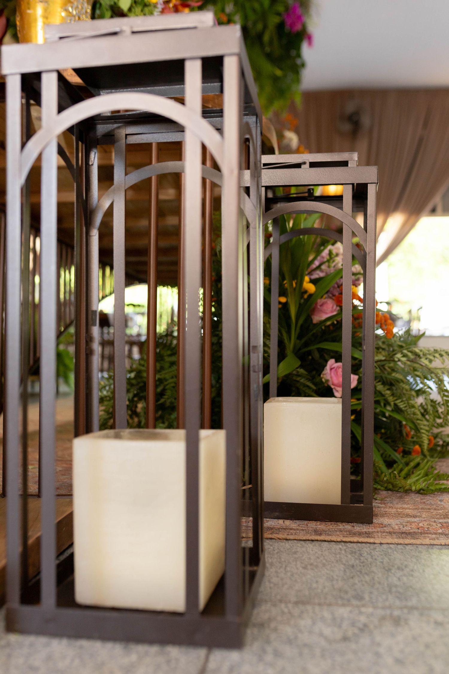 Elegant dark metal lanterns with white pillar candles displayed outdoors among lush green plants and colorful flowers.