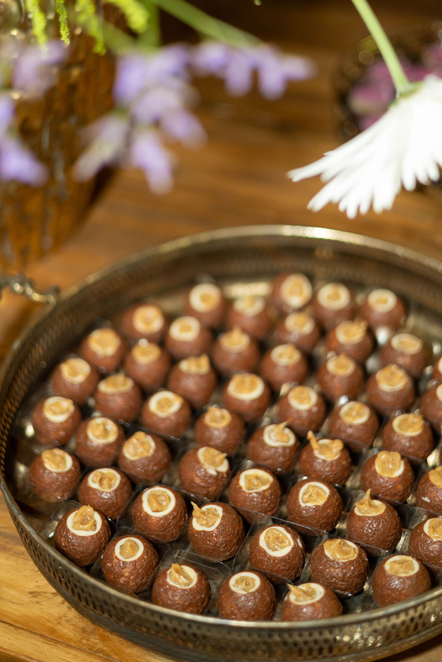 A round silver tray filled with rows of chocolate truffles topped with nuts, displayed on a wooden table with flowers.