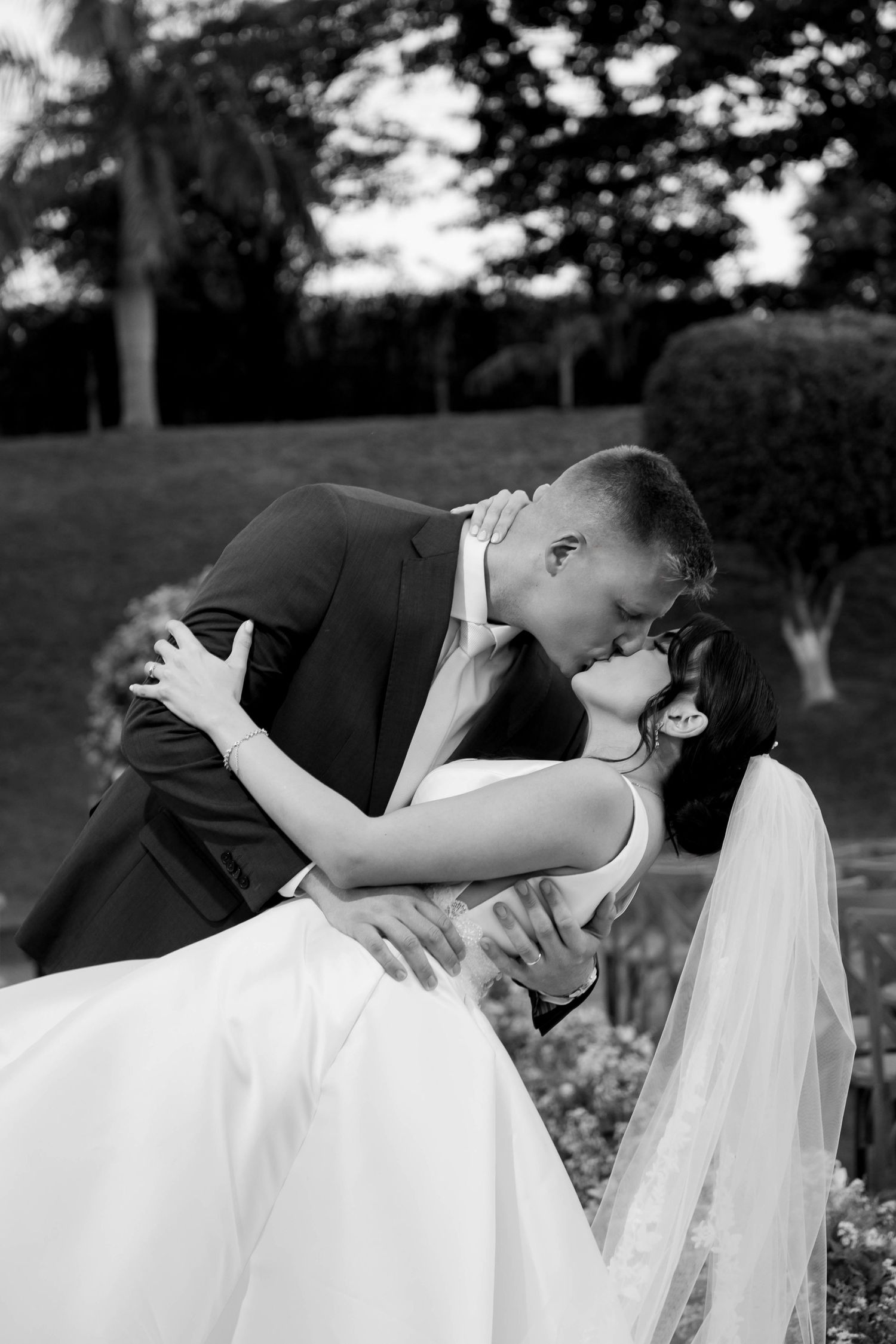 Black and white photo of a groom dipping and kissing his bride in a lush garden on their wedding day.
