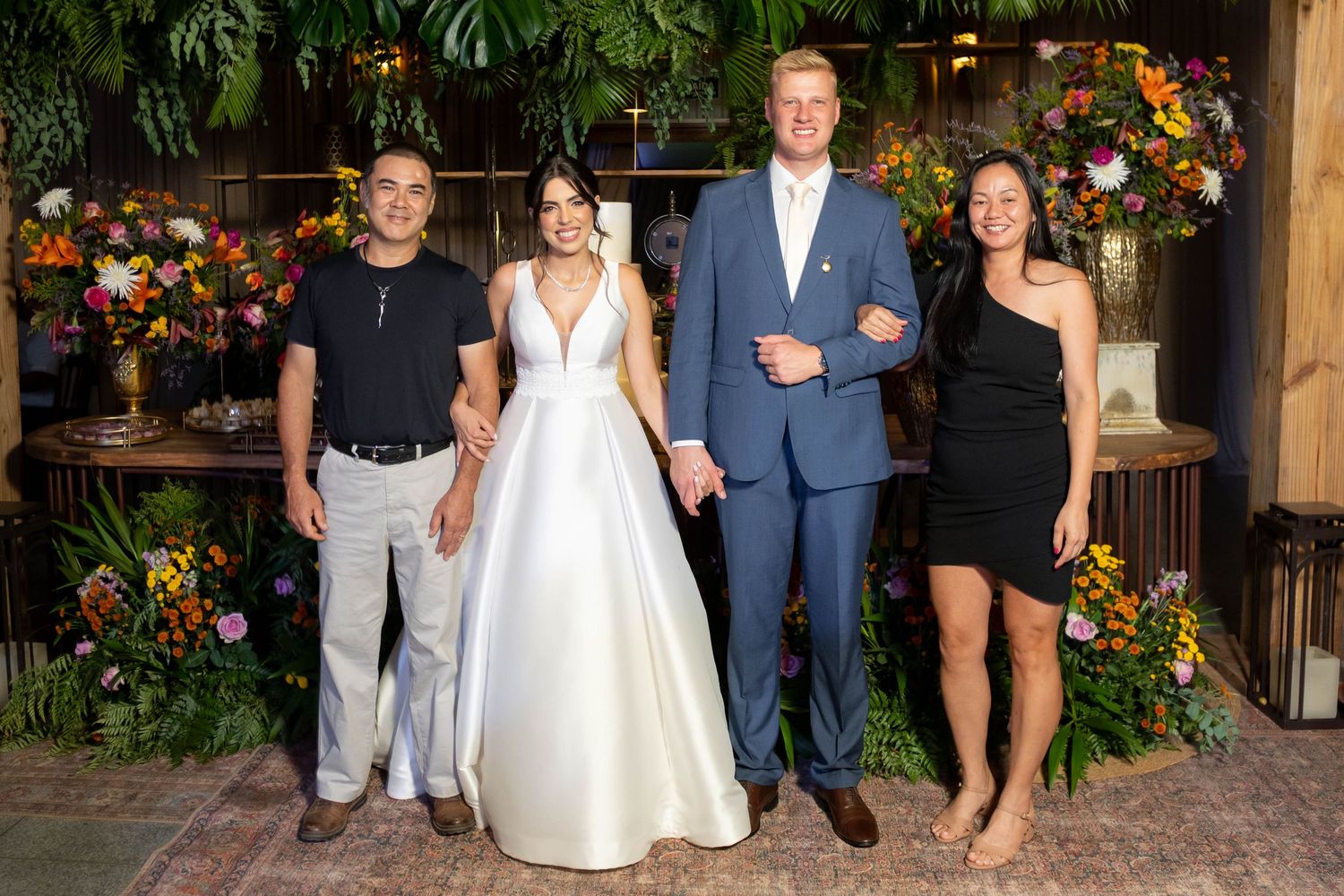 Bride in white gown and groom in blue suit pose with two guests at floral-decorated wedding reception venue.