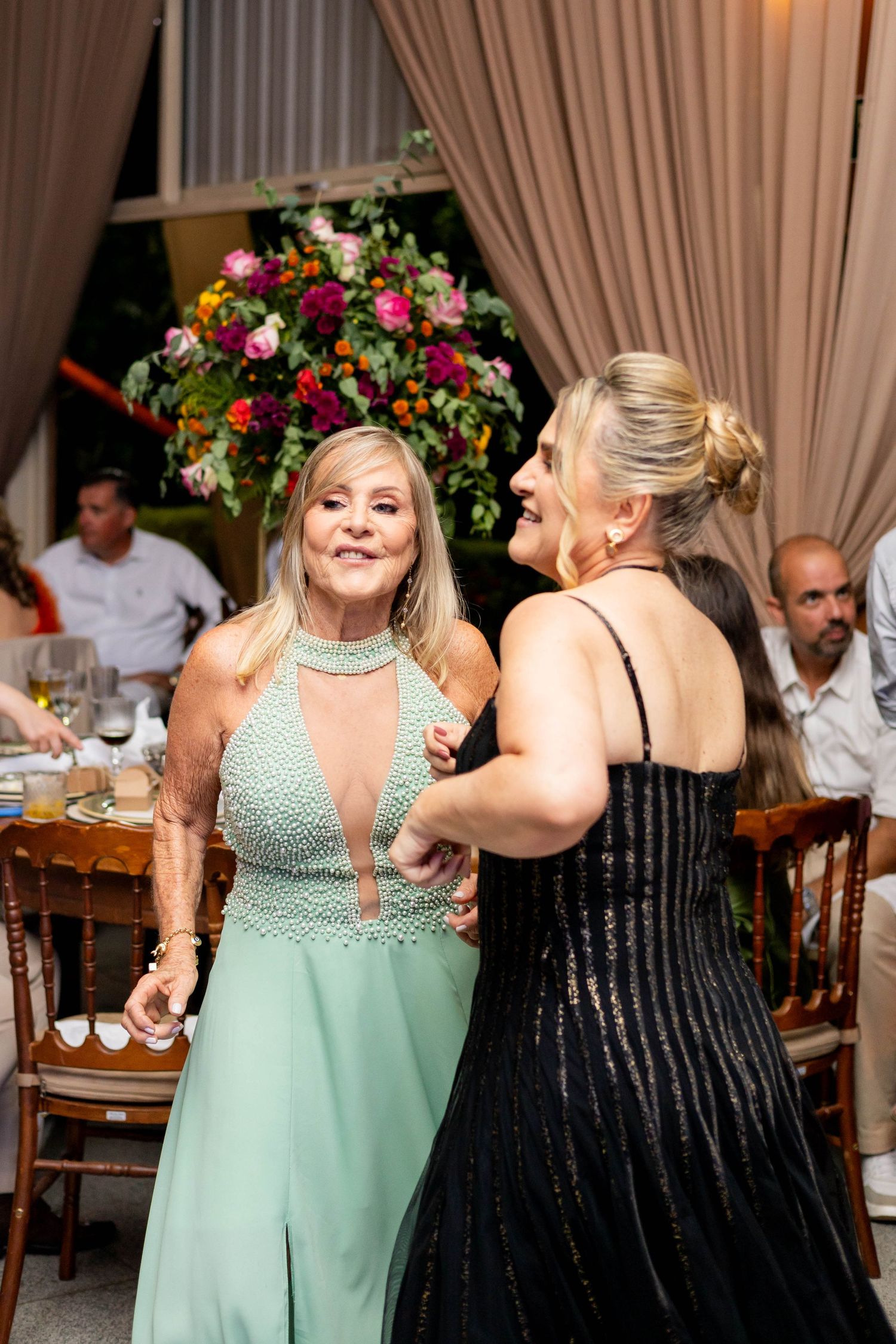 Two women in elegant dresses dancing and laughing at a formal event with floral arrangements in background.