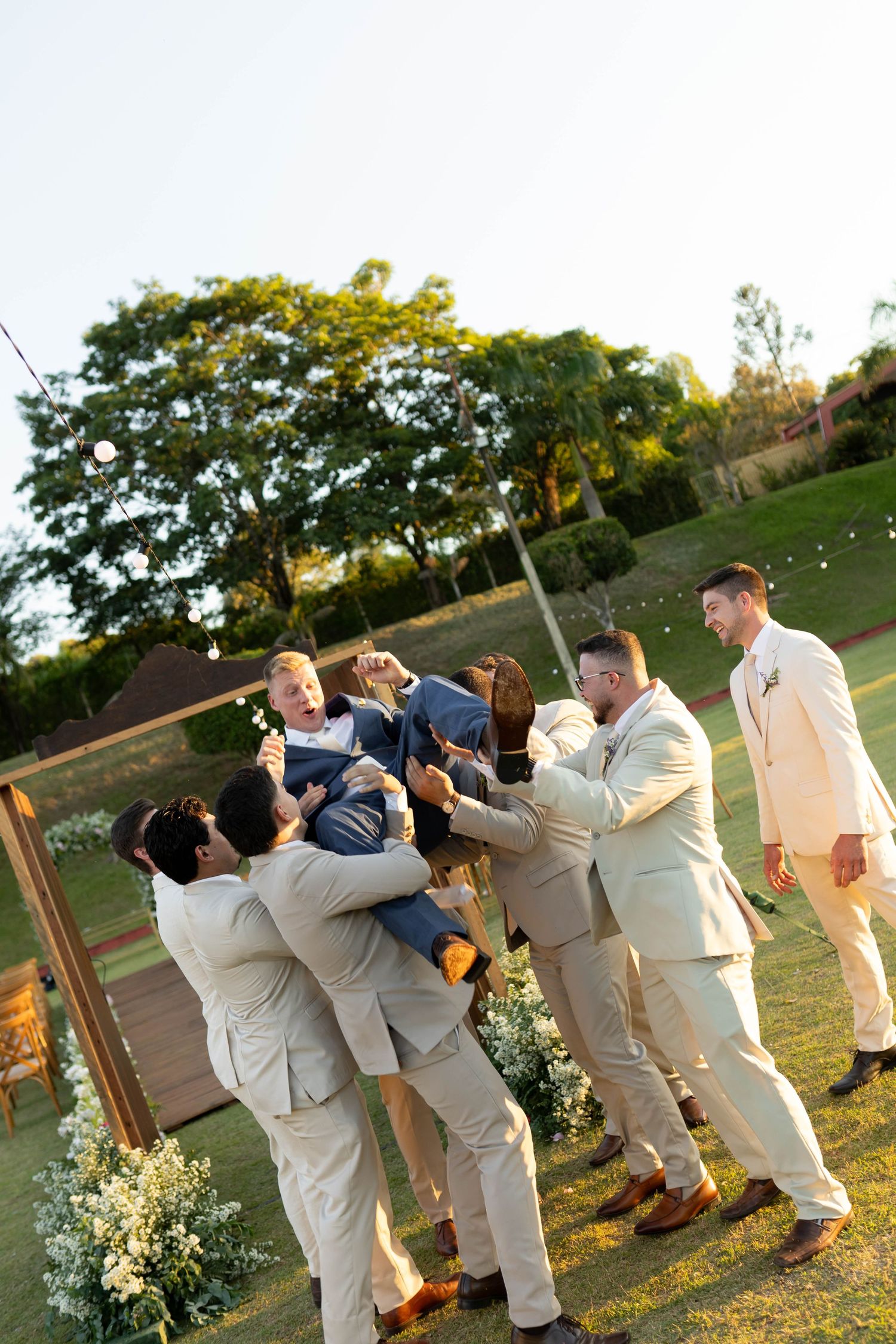 Groomsmen in tan suits lifting groom outdoors at wedding ceremony on green lawn with floral decorations.