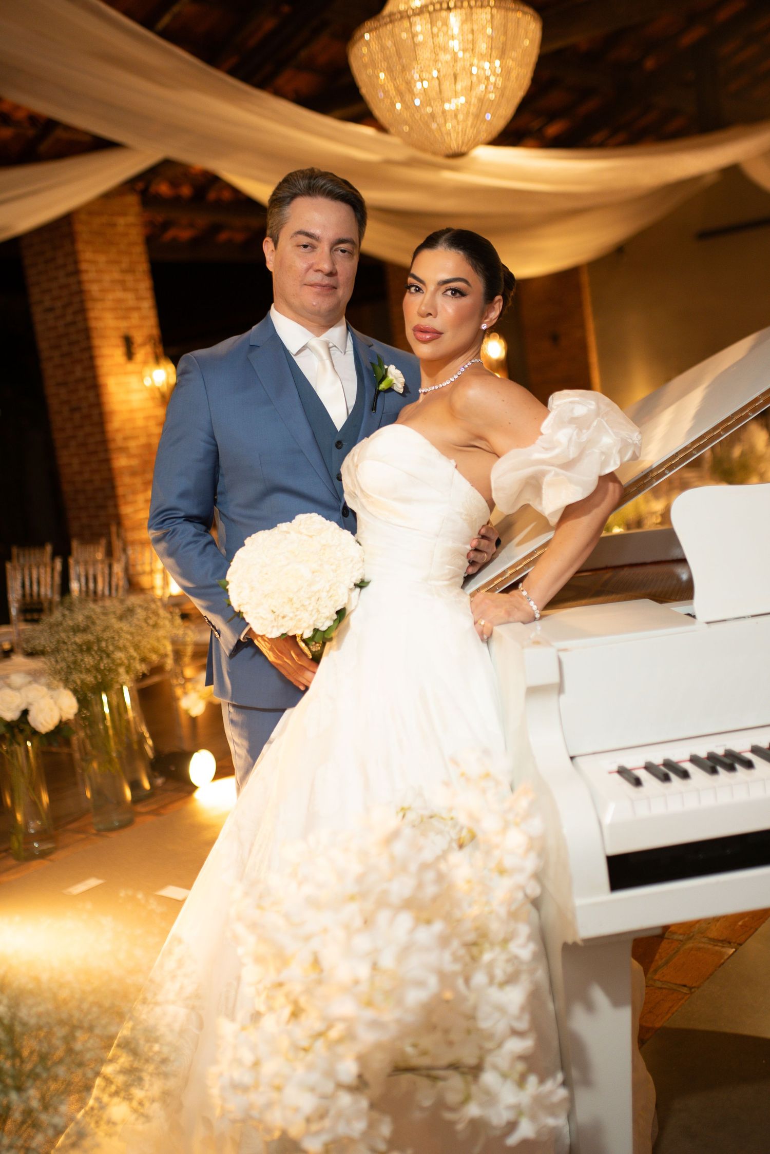 Elegant bride in white gown with bouquet and groom in blue suit pose by white piano at romantic wedding venue.