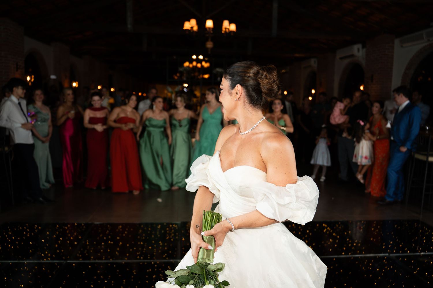 Bride in white off-shoulder gown holds bouquet facing bridesmaids in colorful dresses at elegant wedding reception.