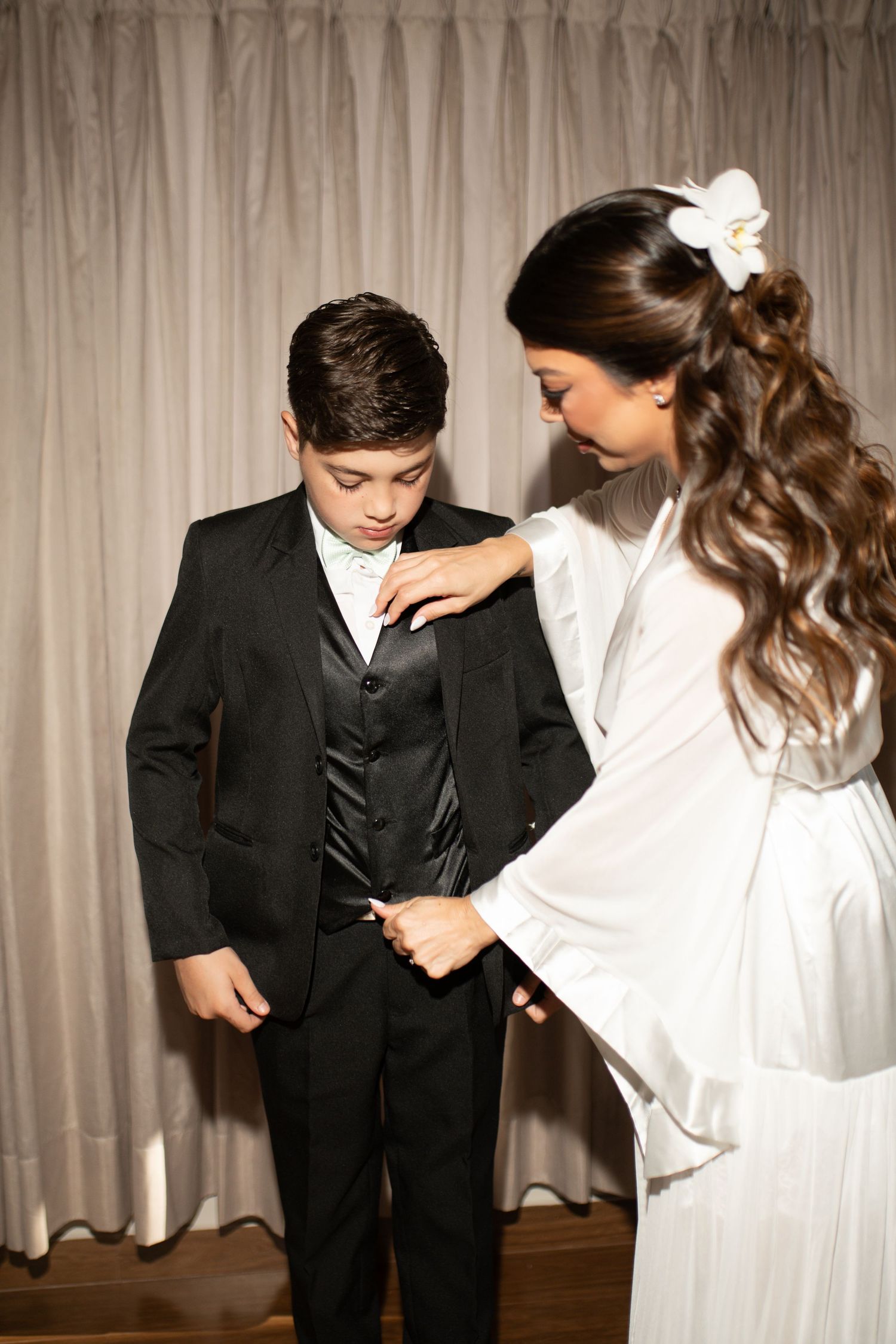 Woman in white dress adjusting a young boy's black tuxedo suit before a formal event.