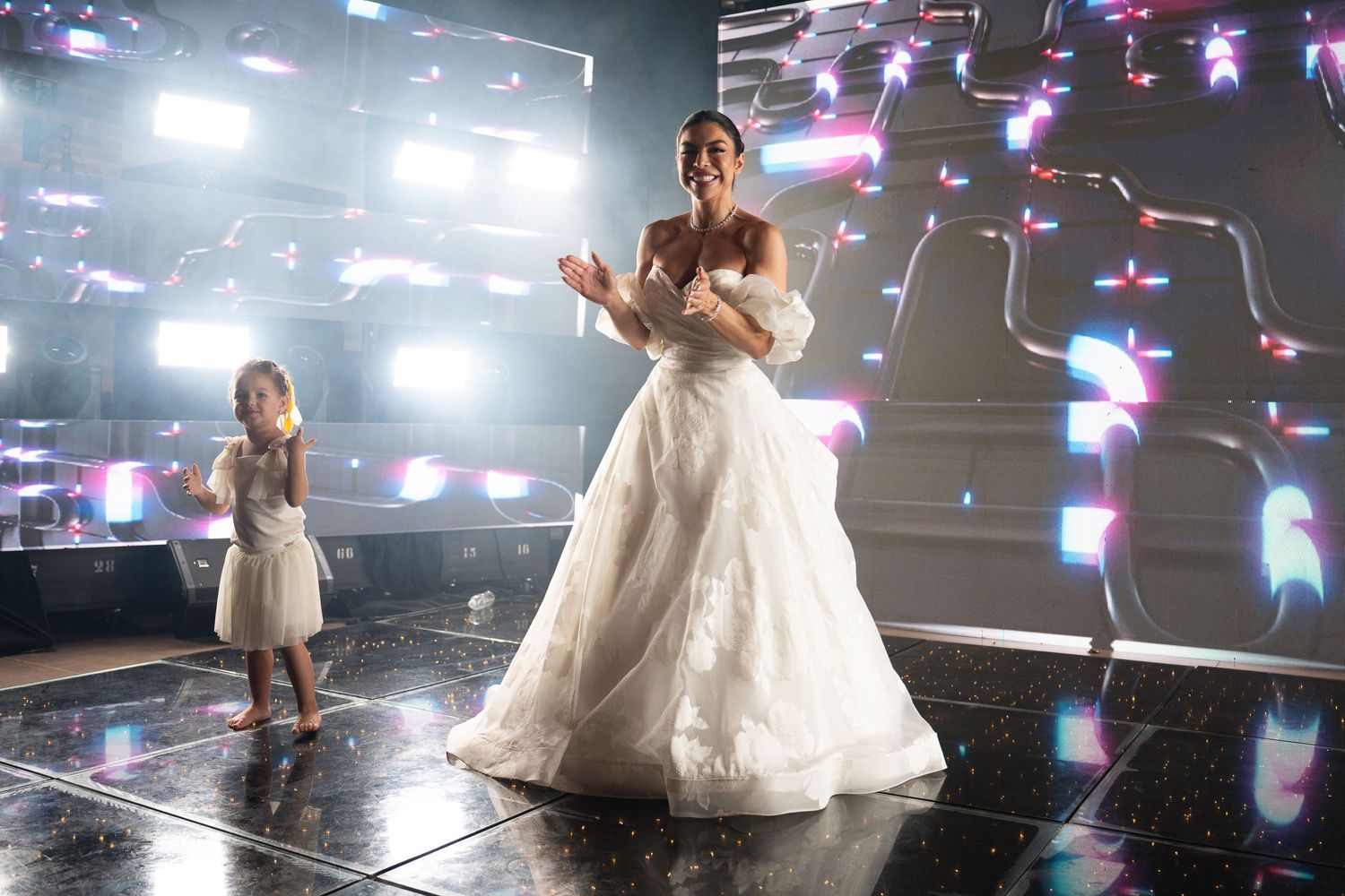 Bride in white ball gown clapping on stage with young flower girl in white dress during wedding reception.