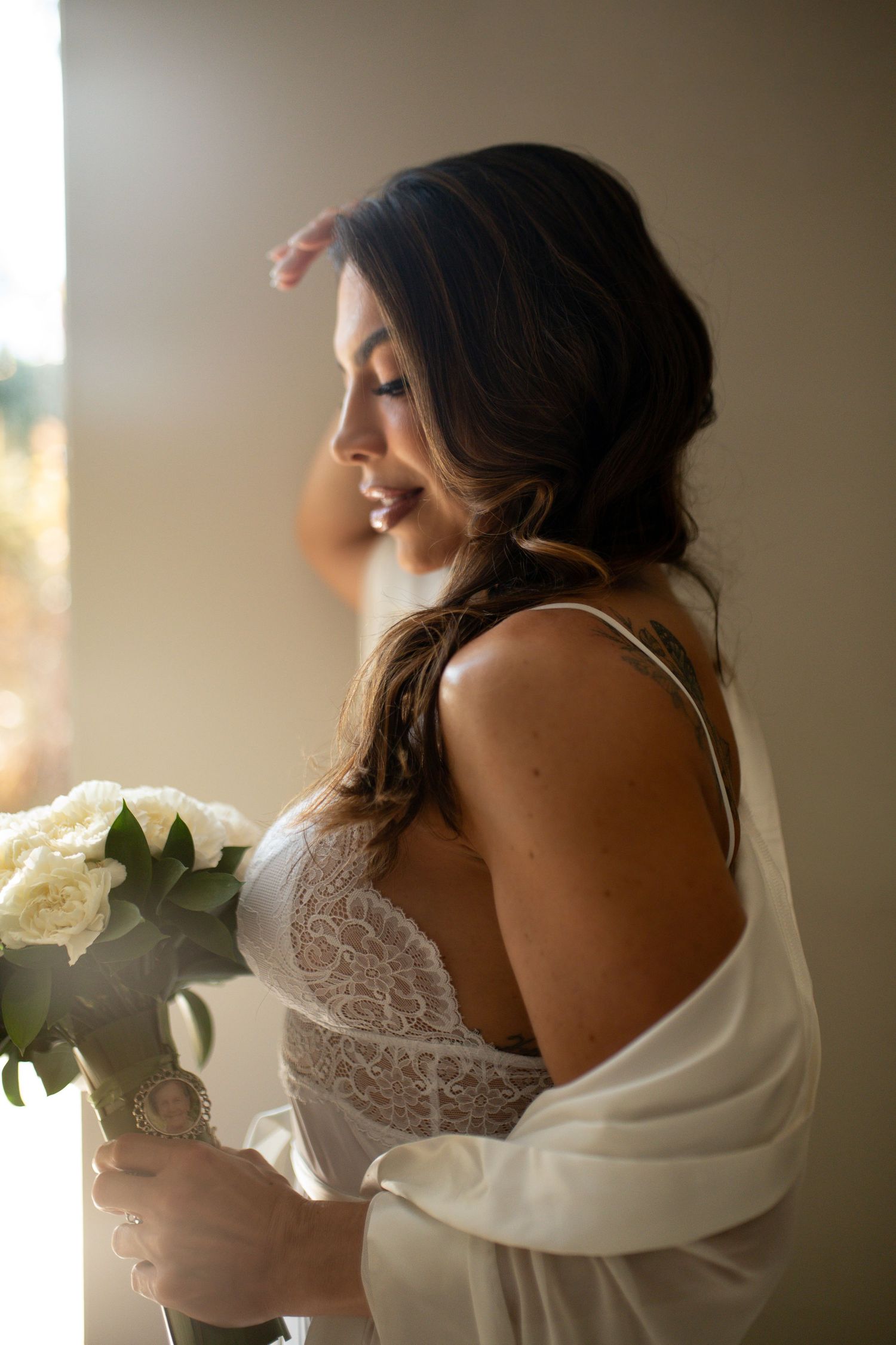 Bride in lace lingerie and white robe holds bouquet of white roses by a sunlit window.