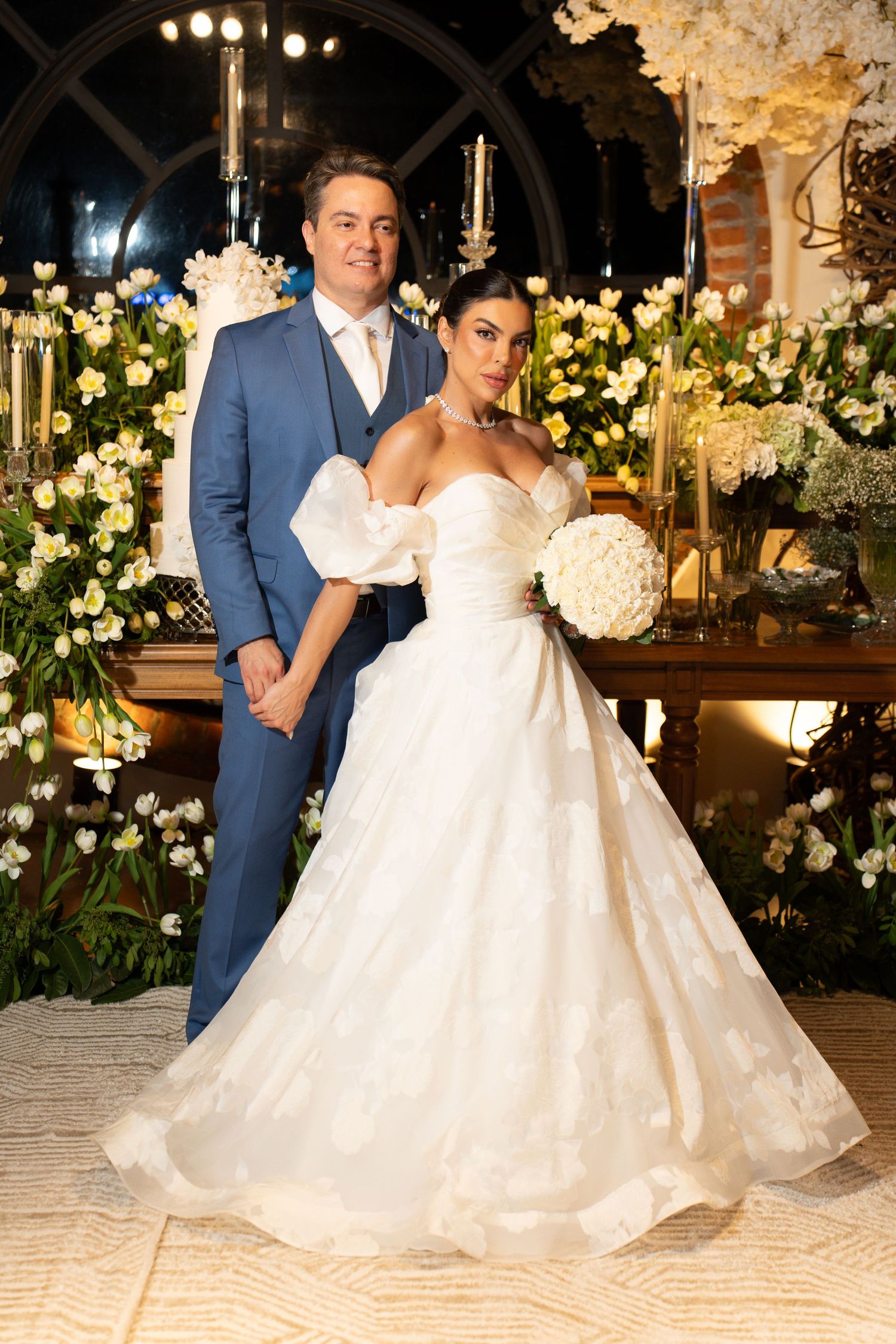Bride in white ball gown with bouquet and groom in blue suit pose together at elegant floral wedding reception.
