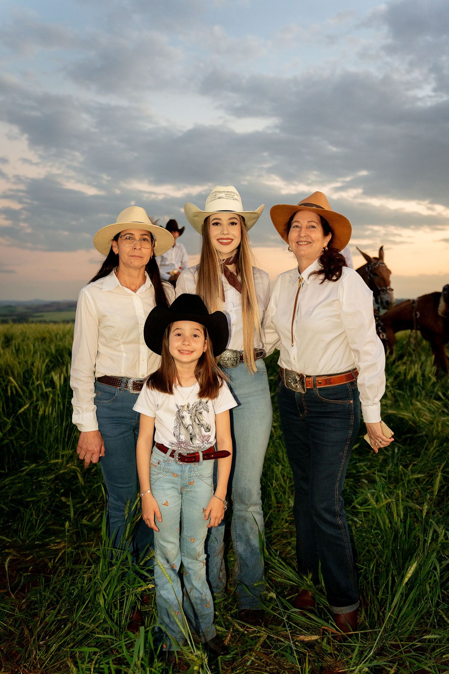 Four females in cowboy hats and jeans pose in a green field at sunset with horses in the background.