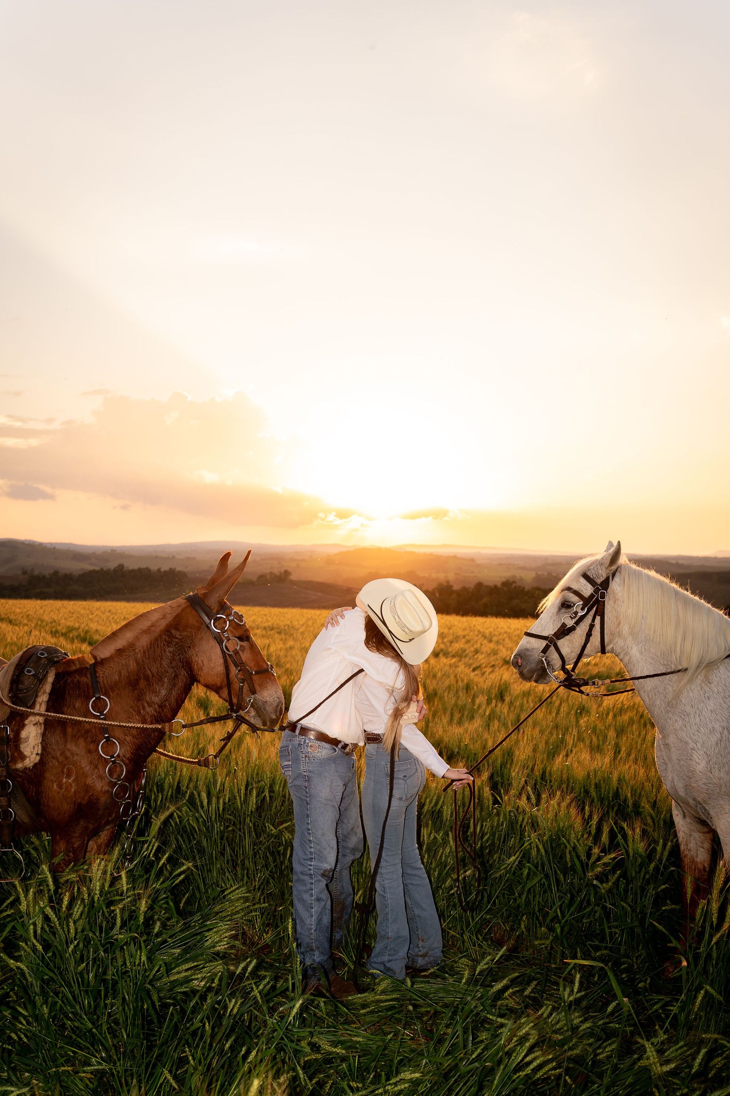 A cowgirl in a white hat and jeans stands between a brown and white horse in a golden wheat field at sunset.
