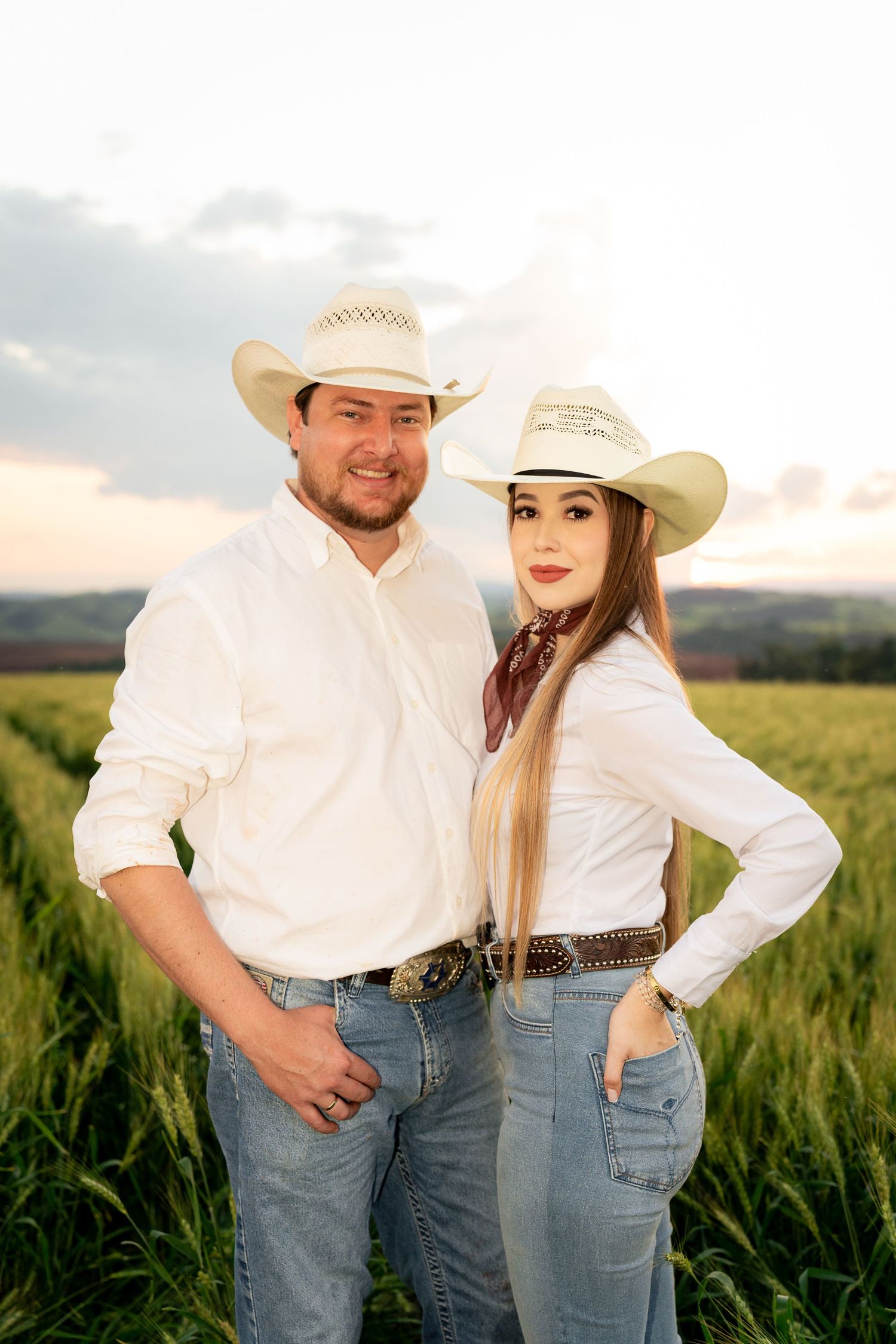 A couple in matching white shirts, cowboy hats, and jeans poses in a green field at sunset.