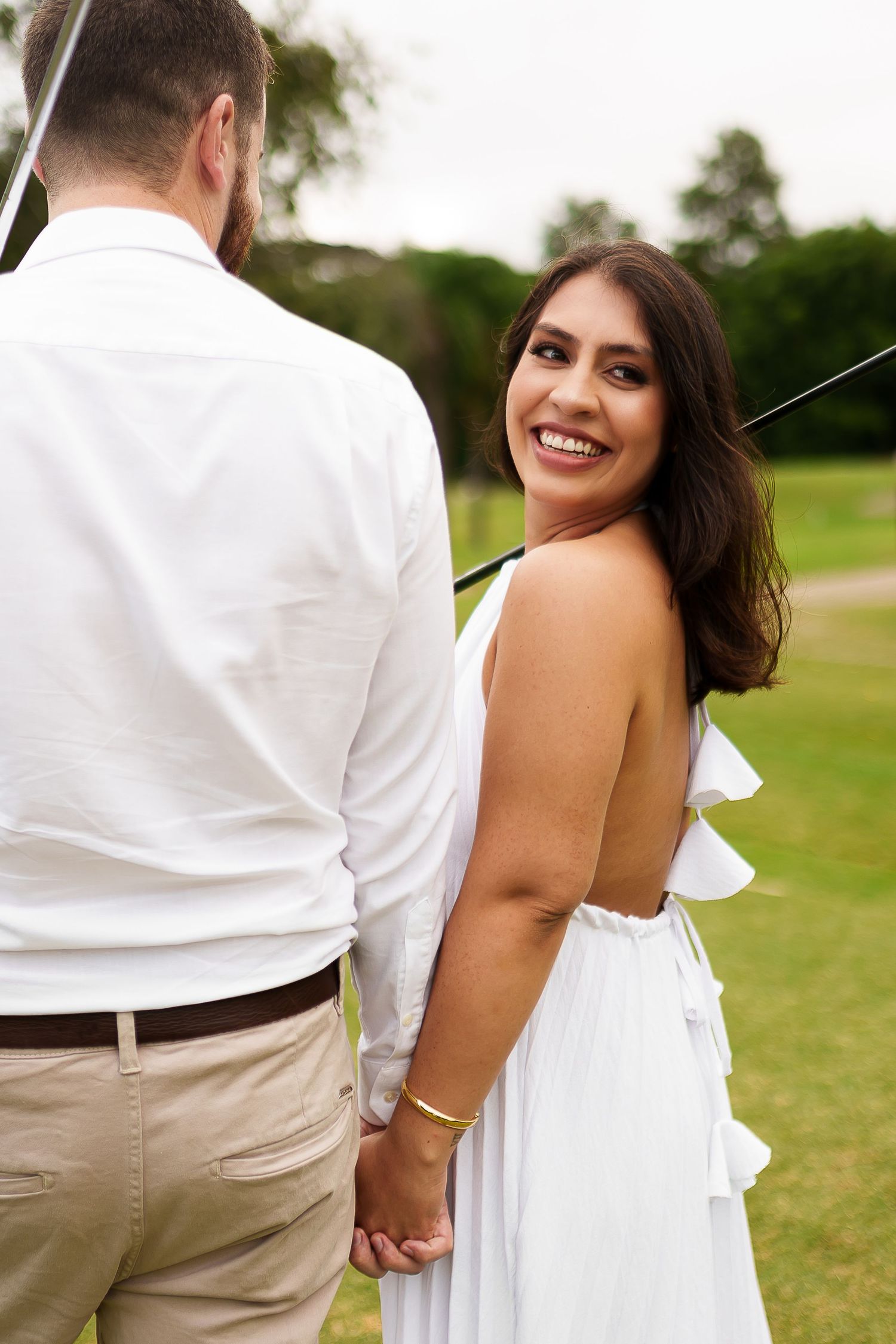A smiling bride in a white dress holds hands with her groom outdoors on a green lawn.
