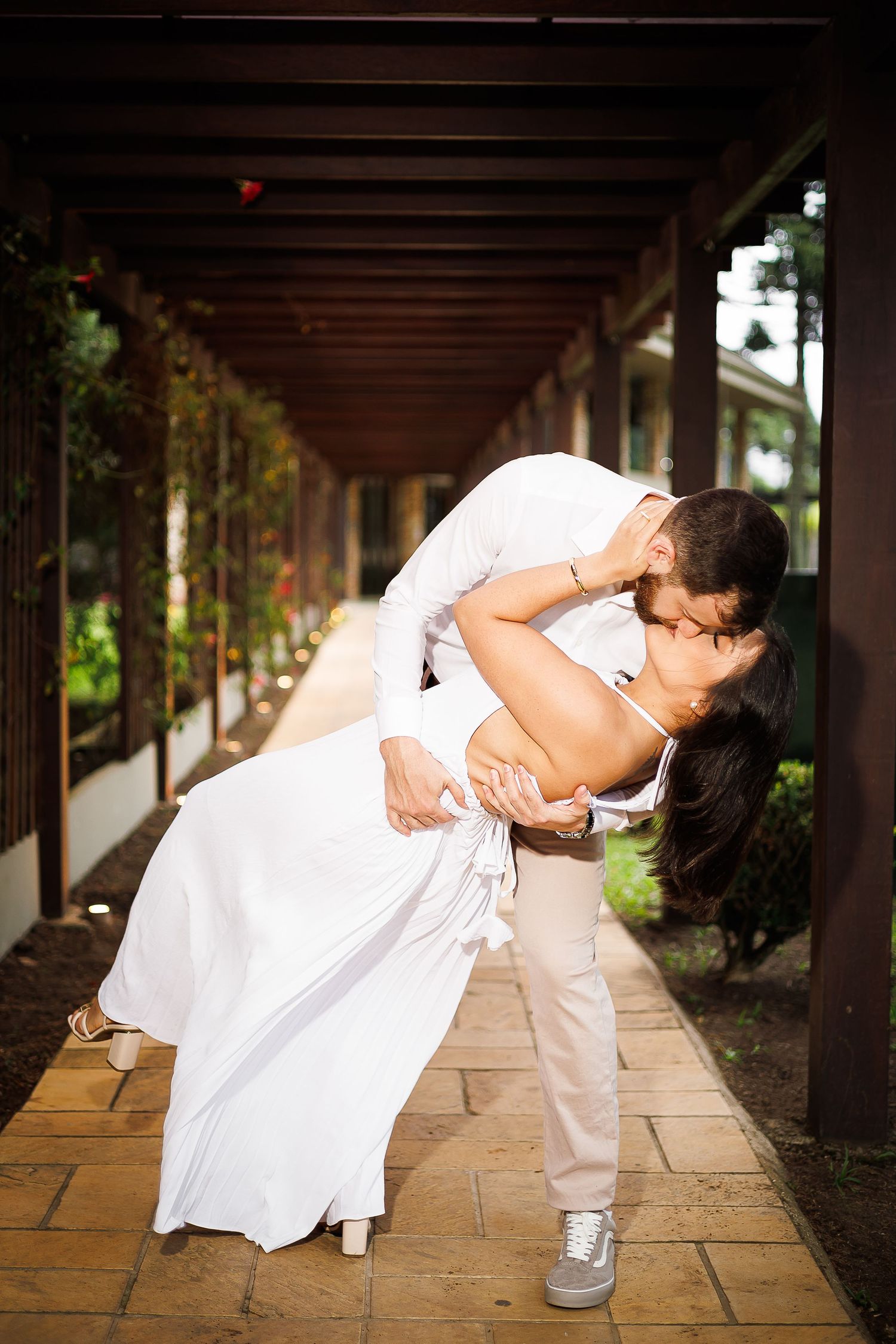 Groom dips bride for a romantic kiss in a sunlit outdoor corridor surrounded by lush greenery on their wedding day.
