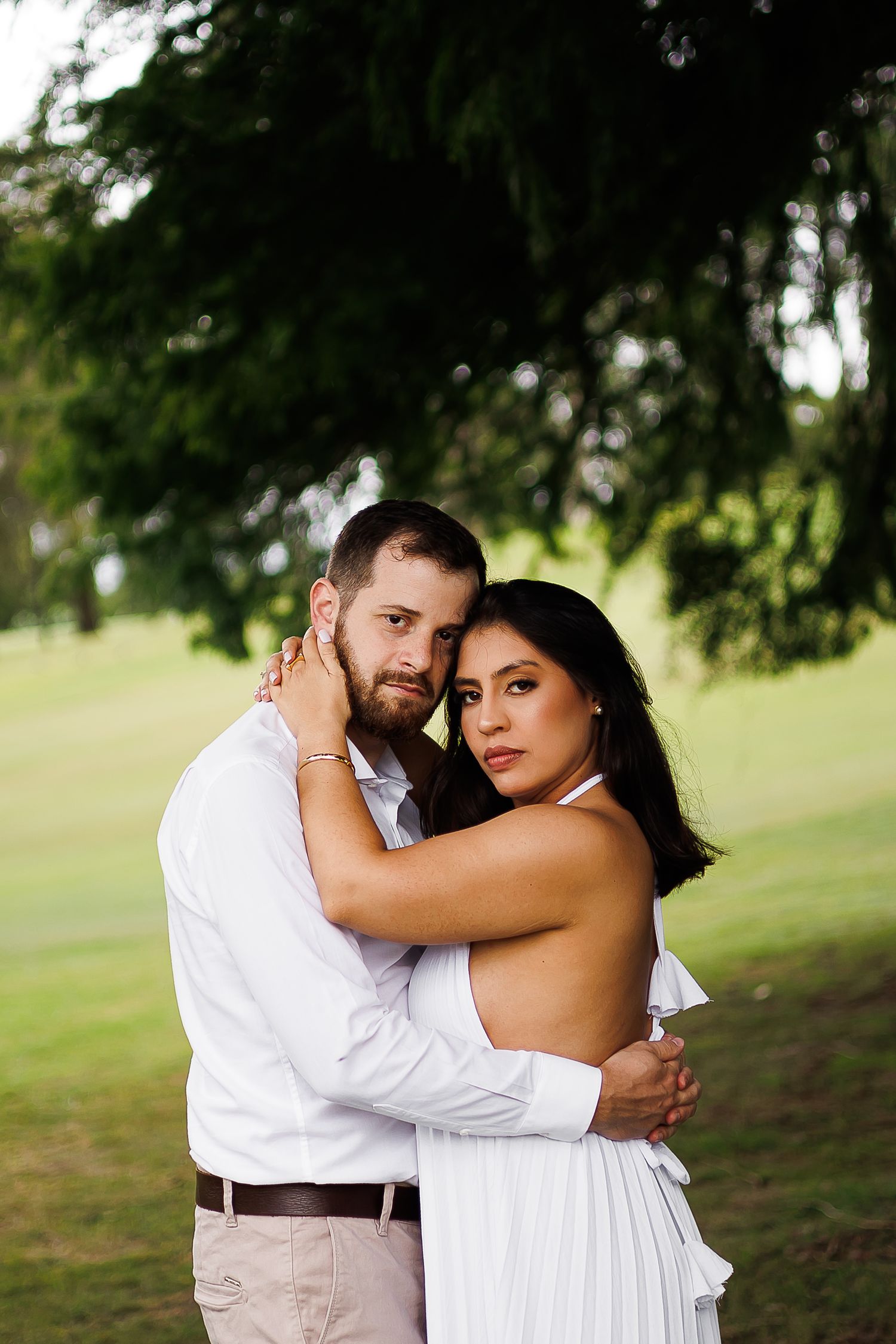Couple embracing outdoors in white outfits under a tree, romantic engagement photo in a lush green park.