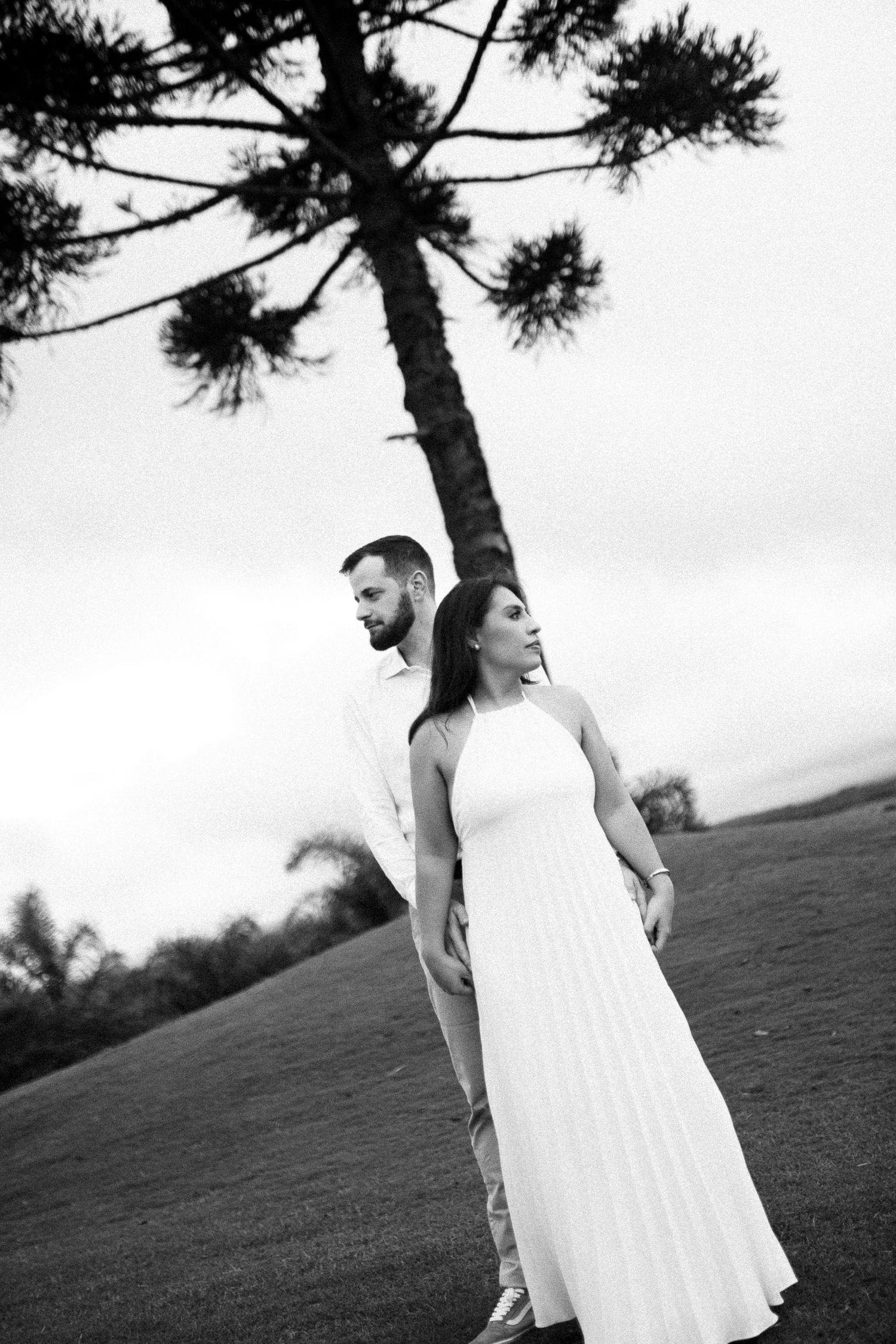 Black and white photo of a couple in white attire standing near a tall tree in an open field.