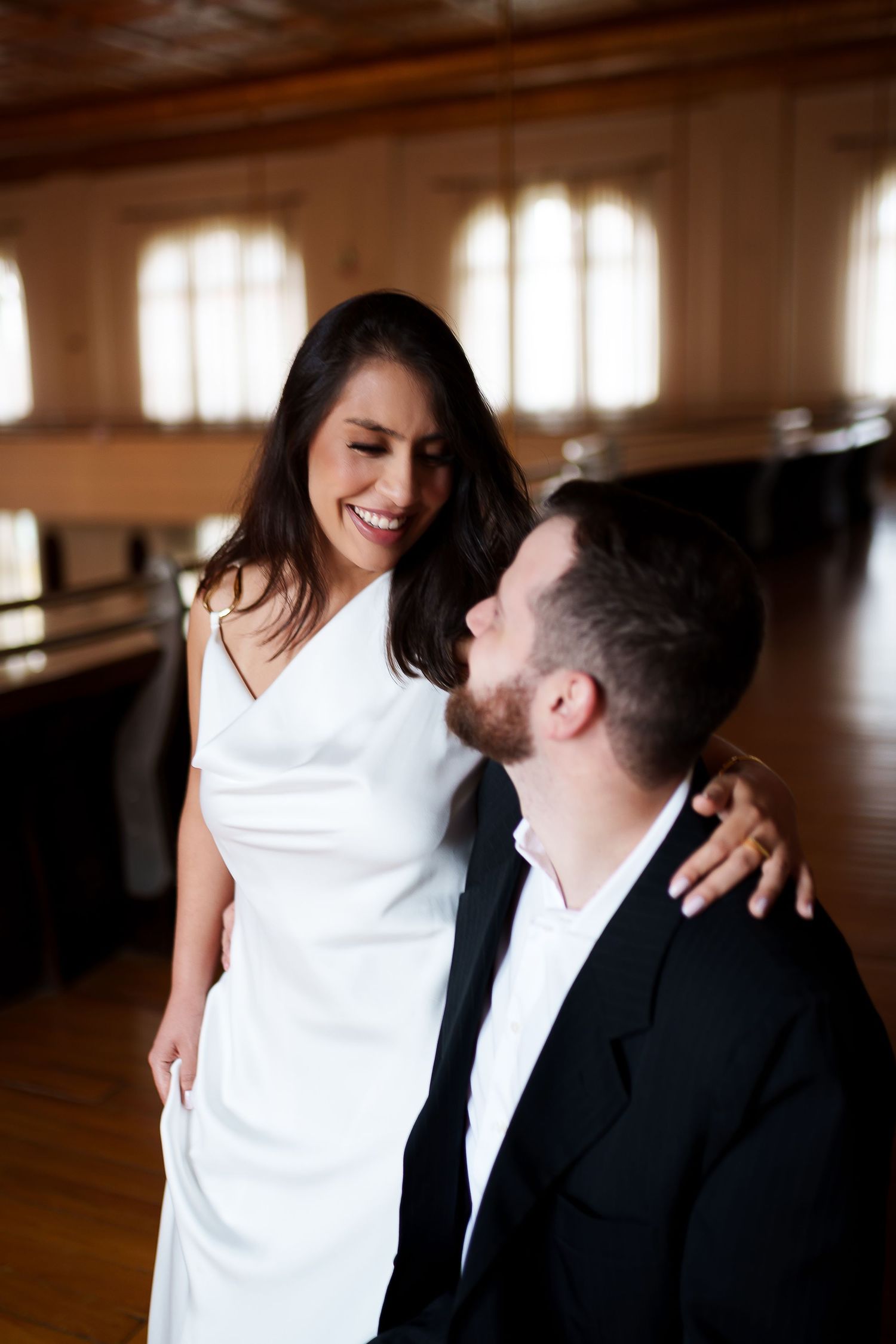 A smiling bride in a white dress embraces a groom in a black suit in an elegant wood-paneled venue.