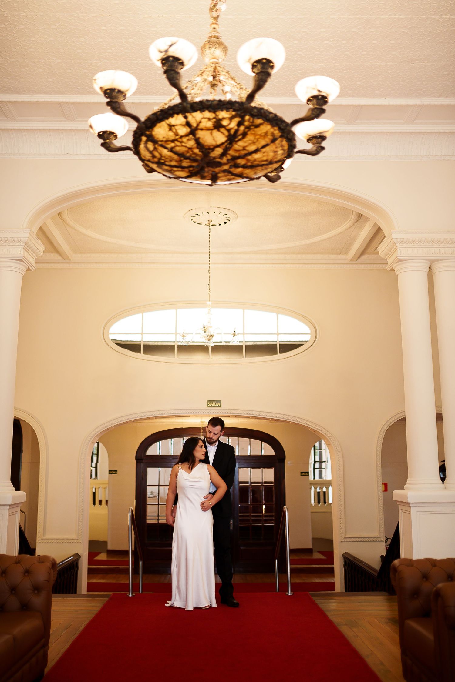 Bride and groom standing on red carpet under ornate chandelier in elegant wedding venue lobby.