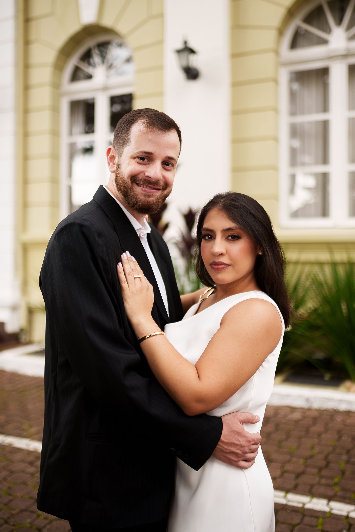 Couple in formal attire embracing and smiling outdoors in front of an elegant building.