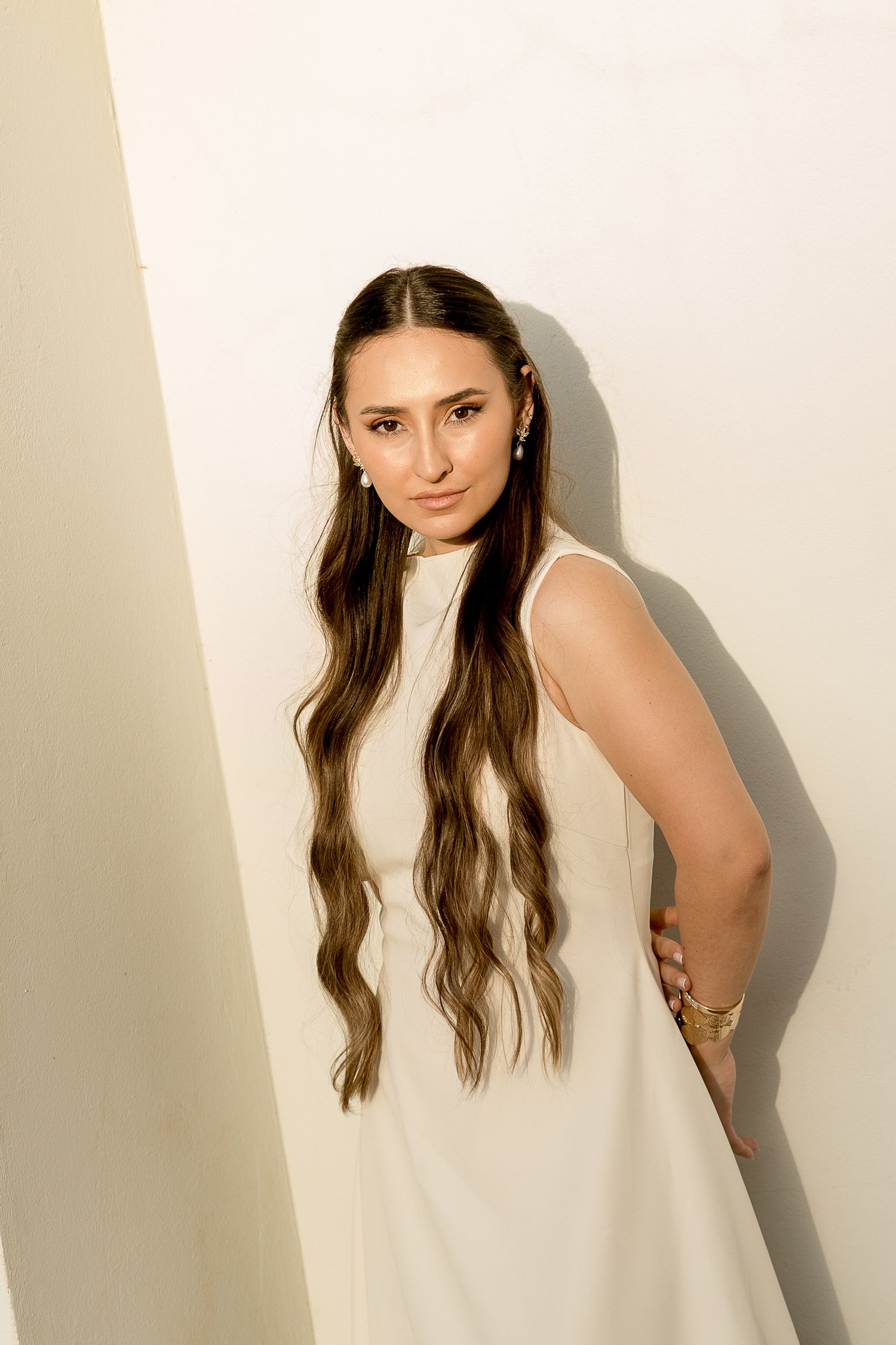 Young woman in white sleeveless dress with long wavy hair poses against white wall wearing gold bracelet.