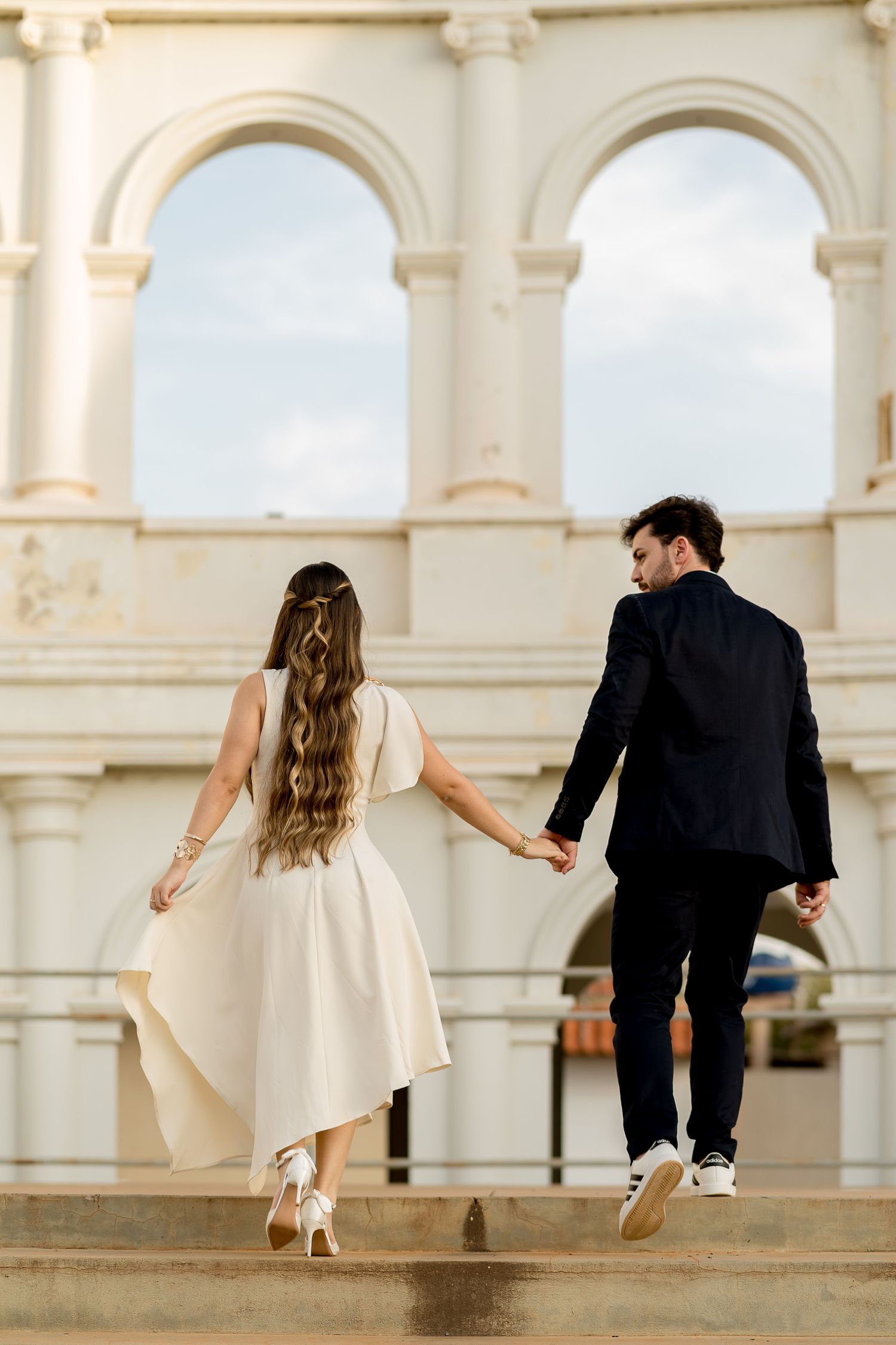 A couple in wedding attire holds hands walking up steps toward elegant arched building exterior.